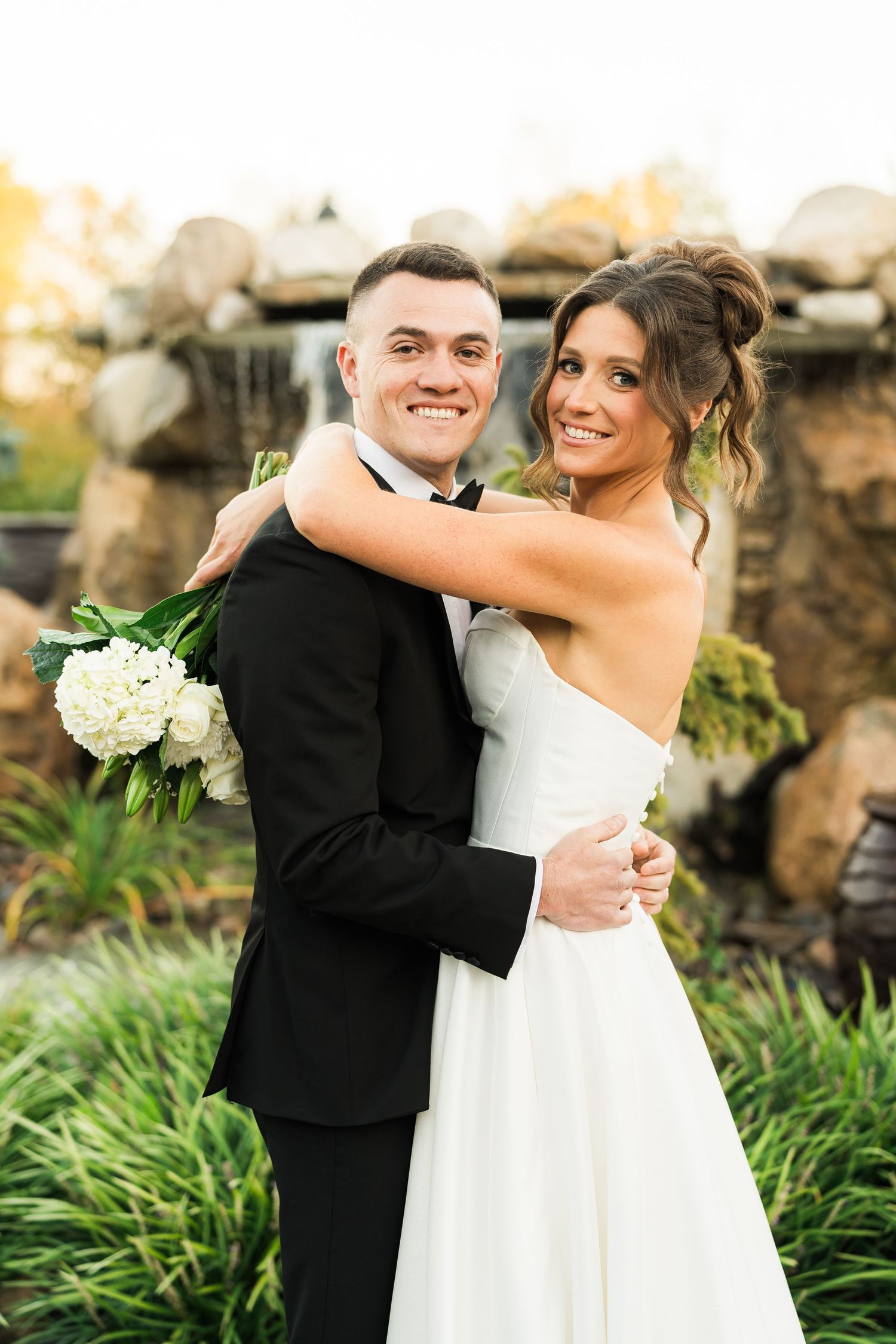 A joyful wedding portrait of a couple embracing outdoors with white flowers and natural scenery.
