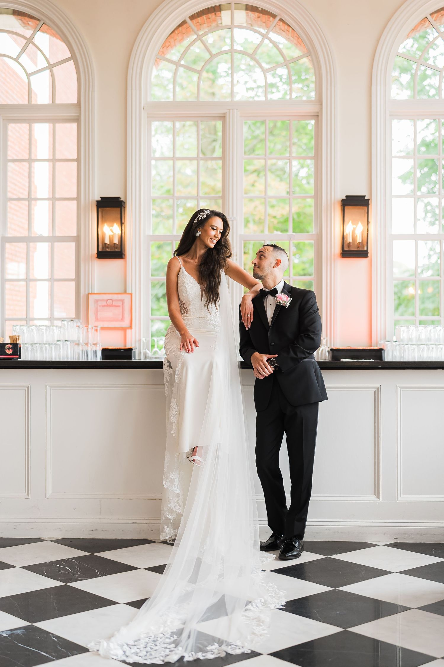 Elegant couple posing in front of arched windows on black and white checkered floor.