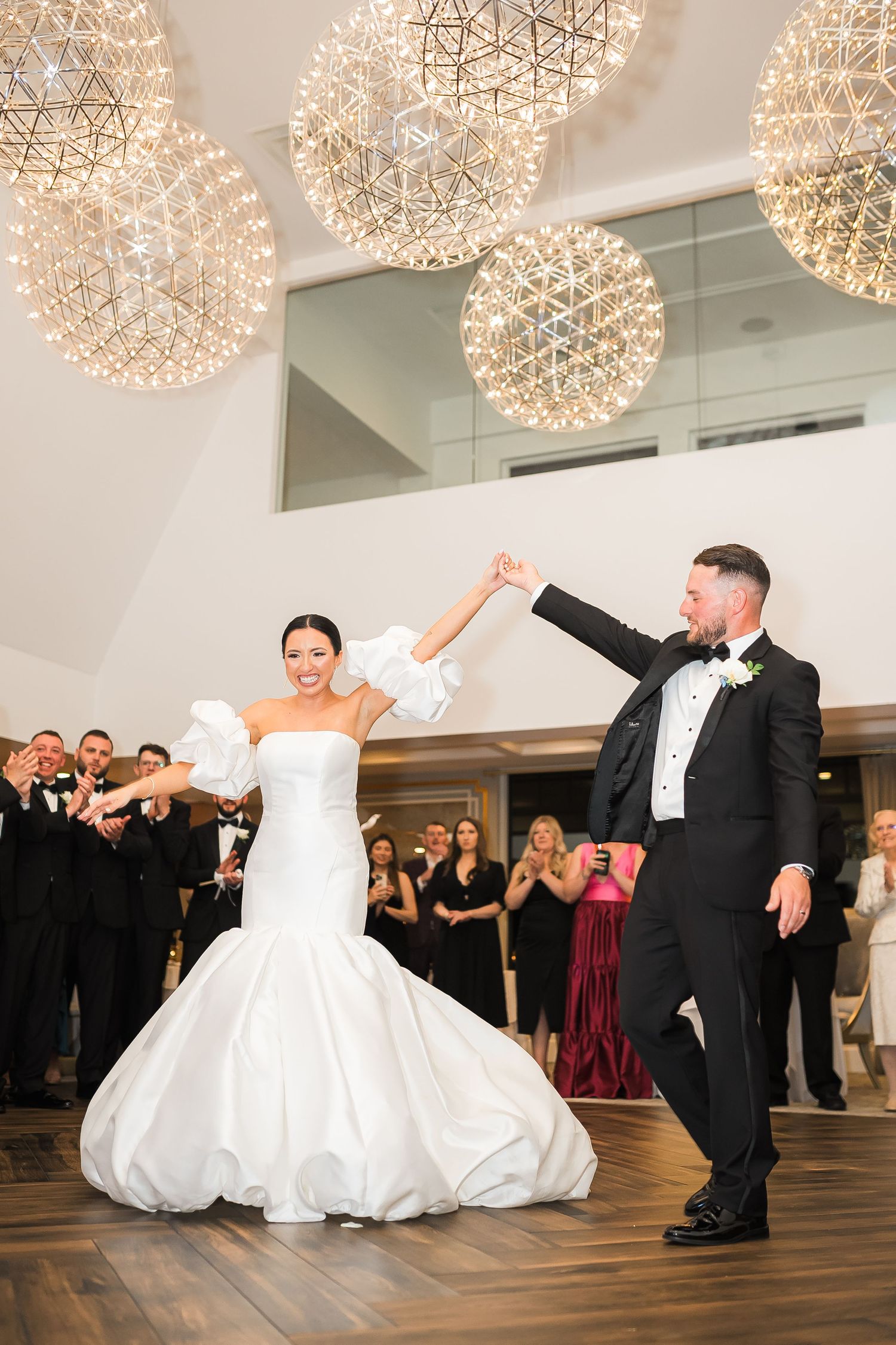 Newlyweds share their first dance under sparkling globe lights while wedding guests cheer them on.
