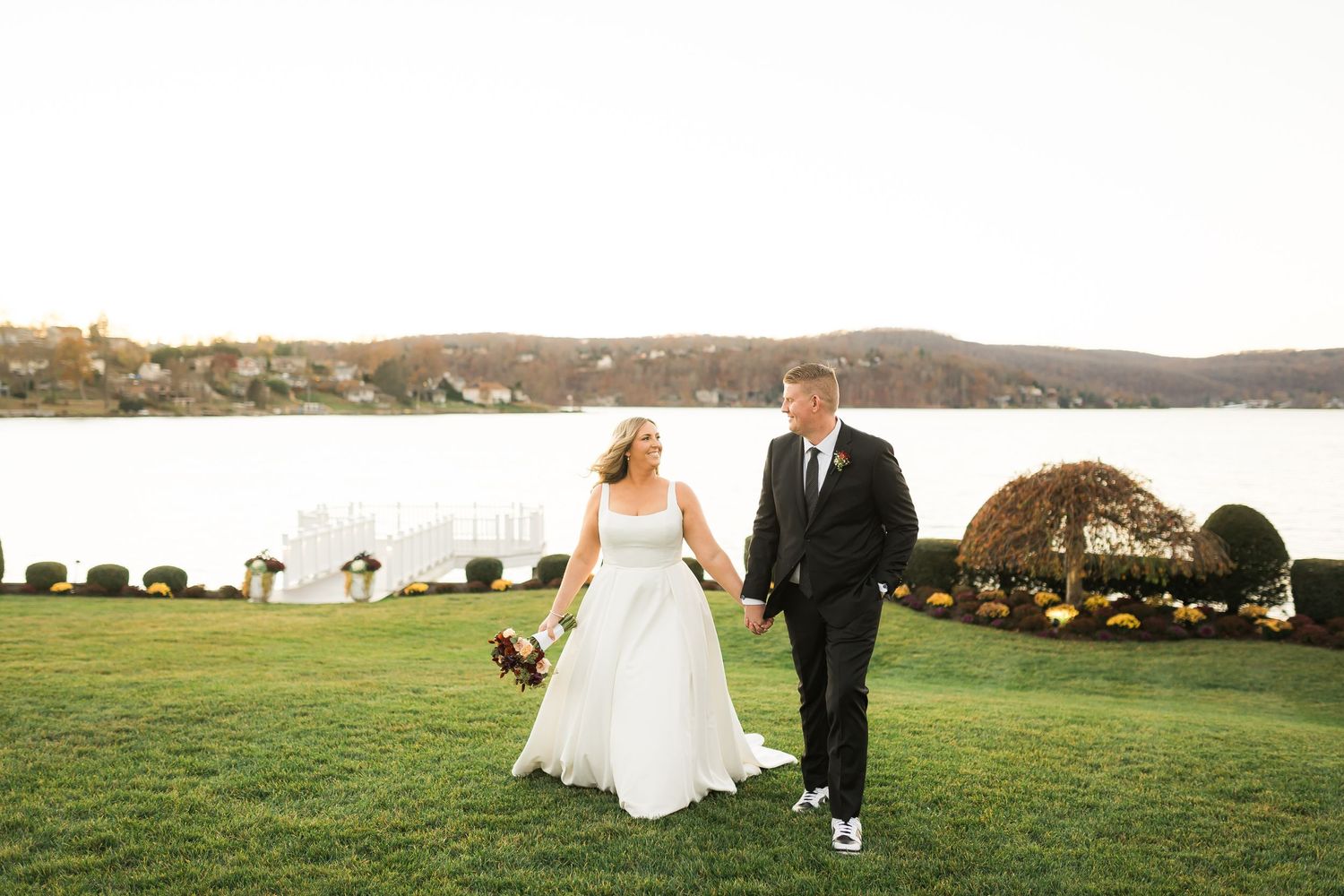 A couple walks hand in hand on a waterfront lawn during their wedding day with water and hills in background.