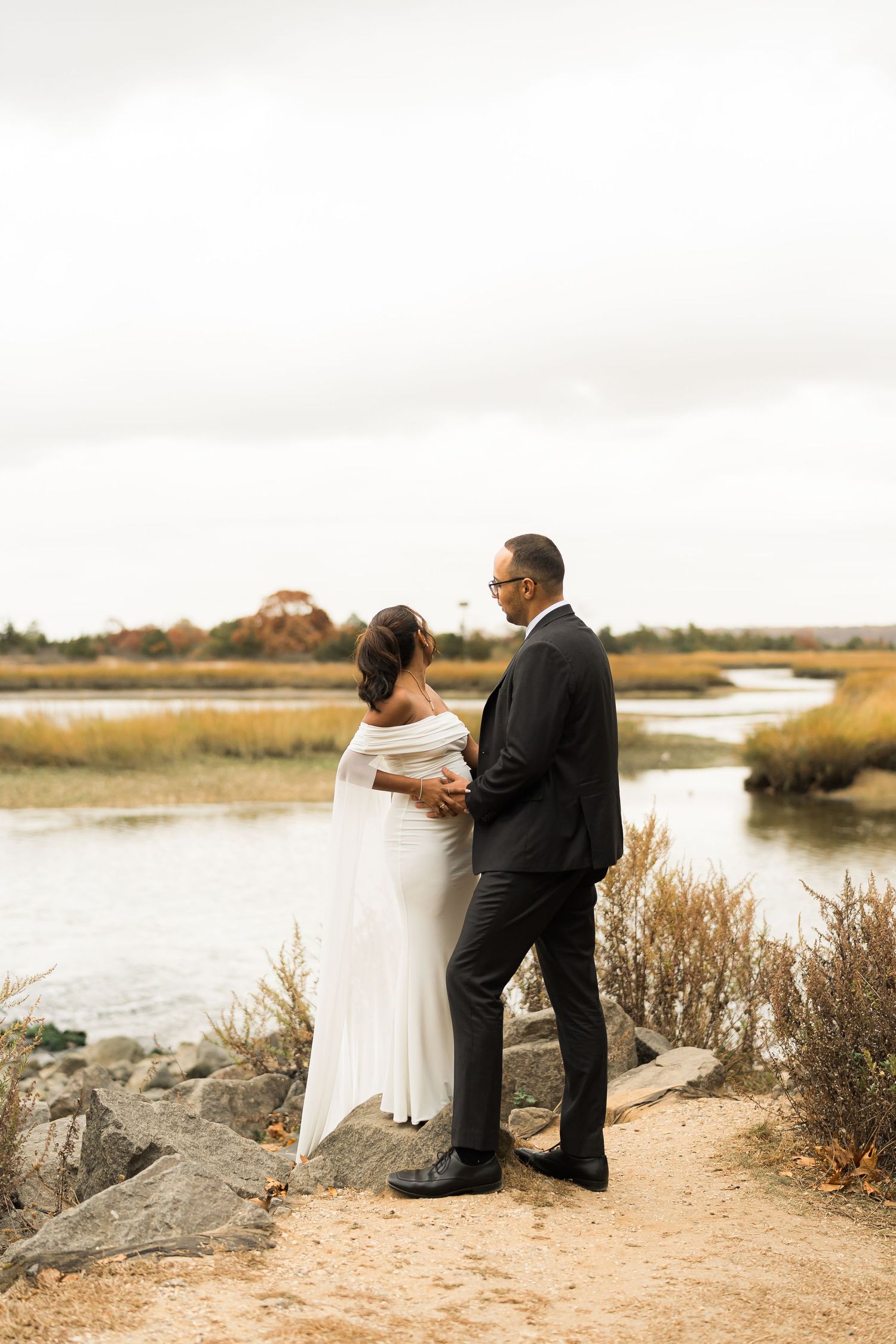 Couple in wedding attire standing together overlooking a scenic marsh landscape.