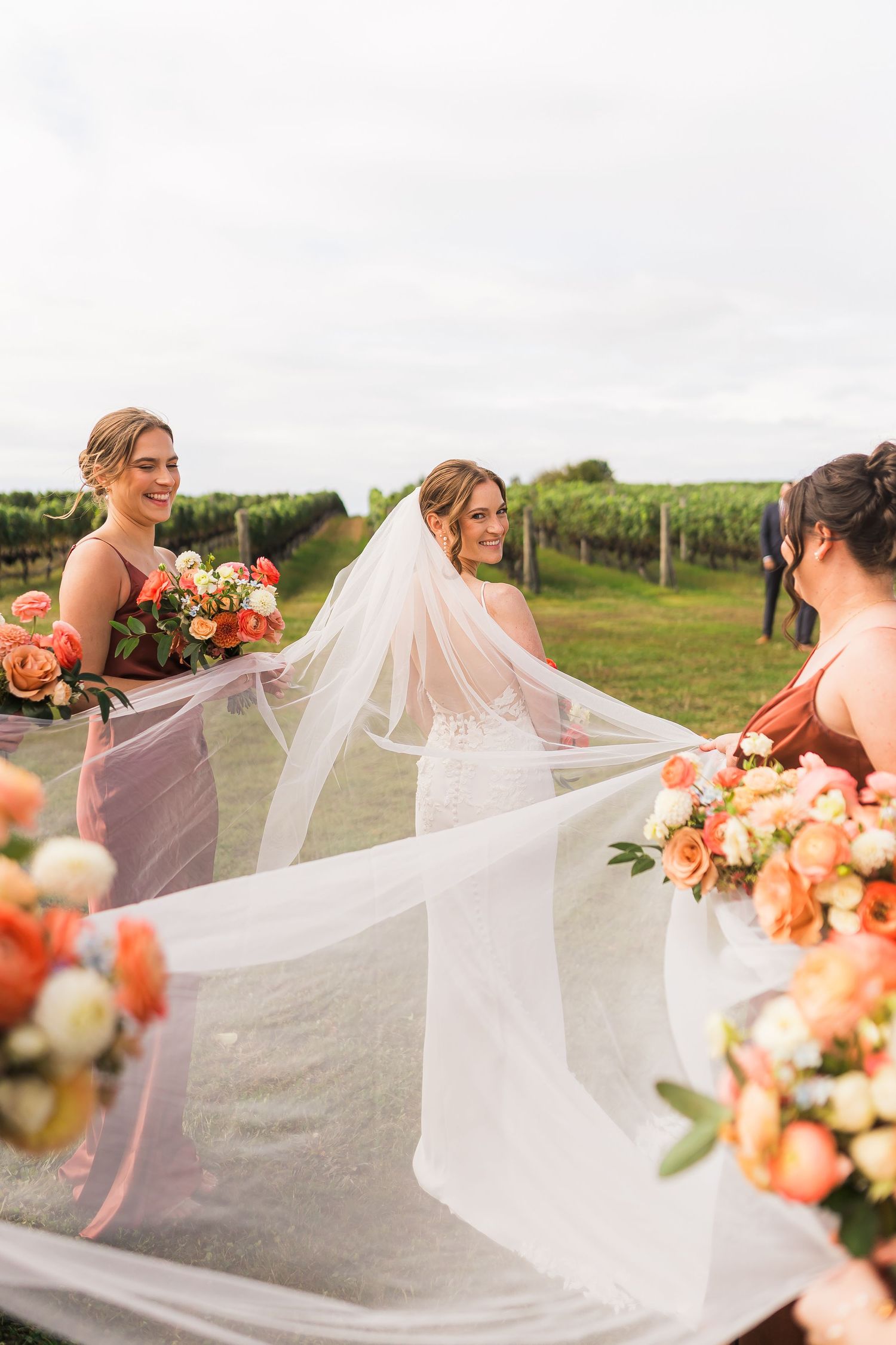 A bride in a flowing veil stands in a vineyard with bridesmaids holding coral and cream floral bouquets.