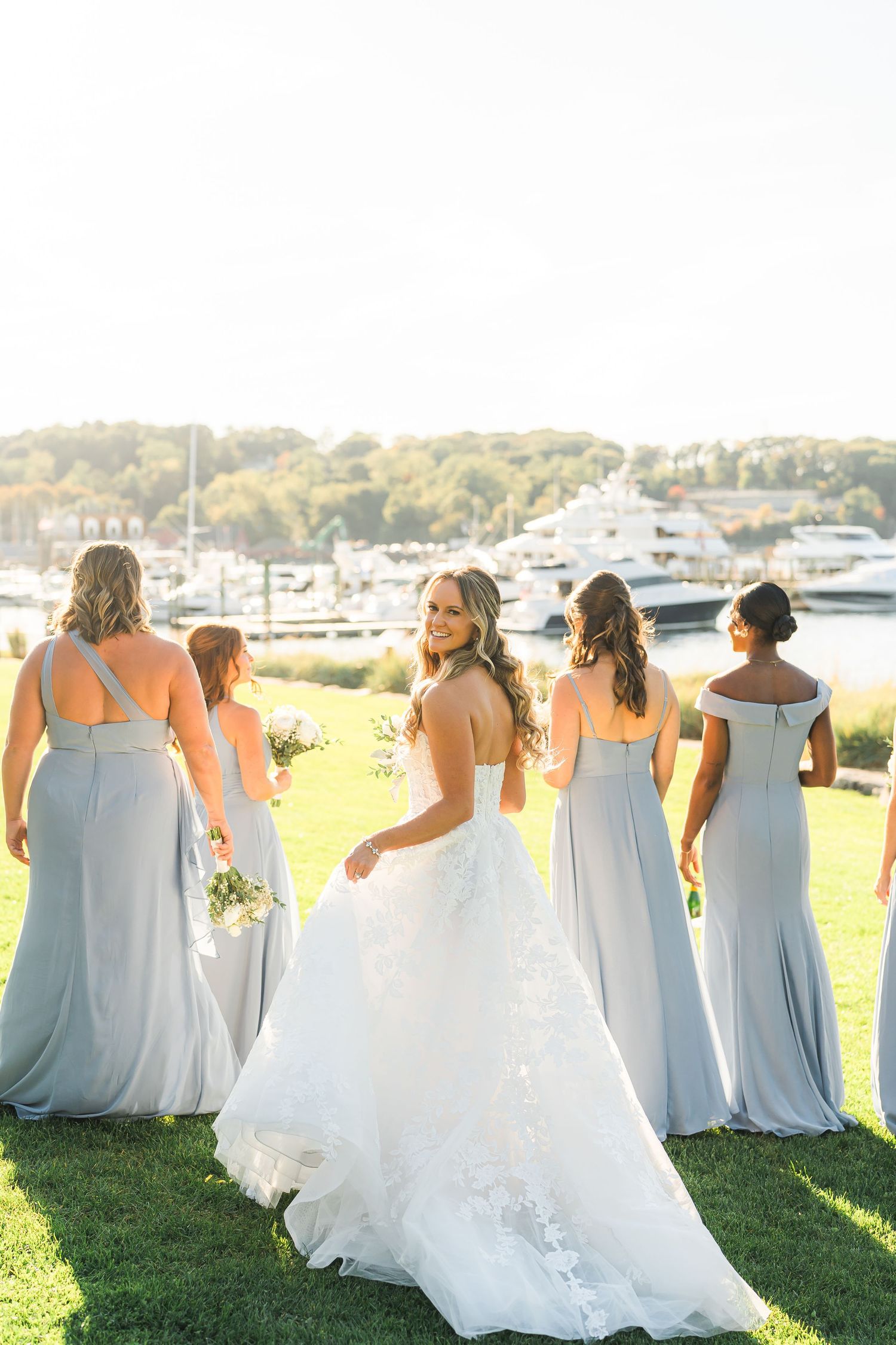 Wedding party in light blue dresses pose near marina at sunset.