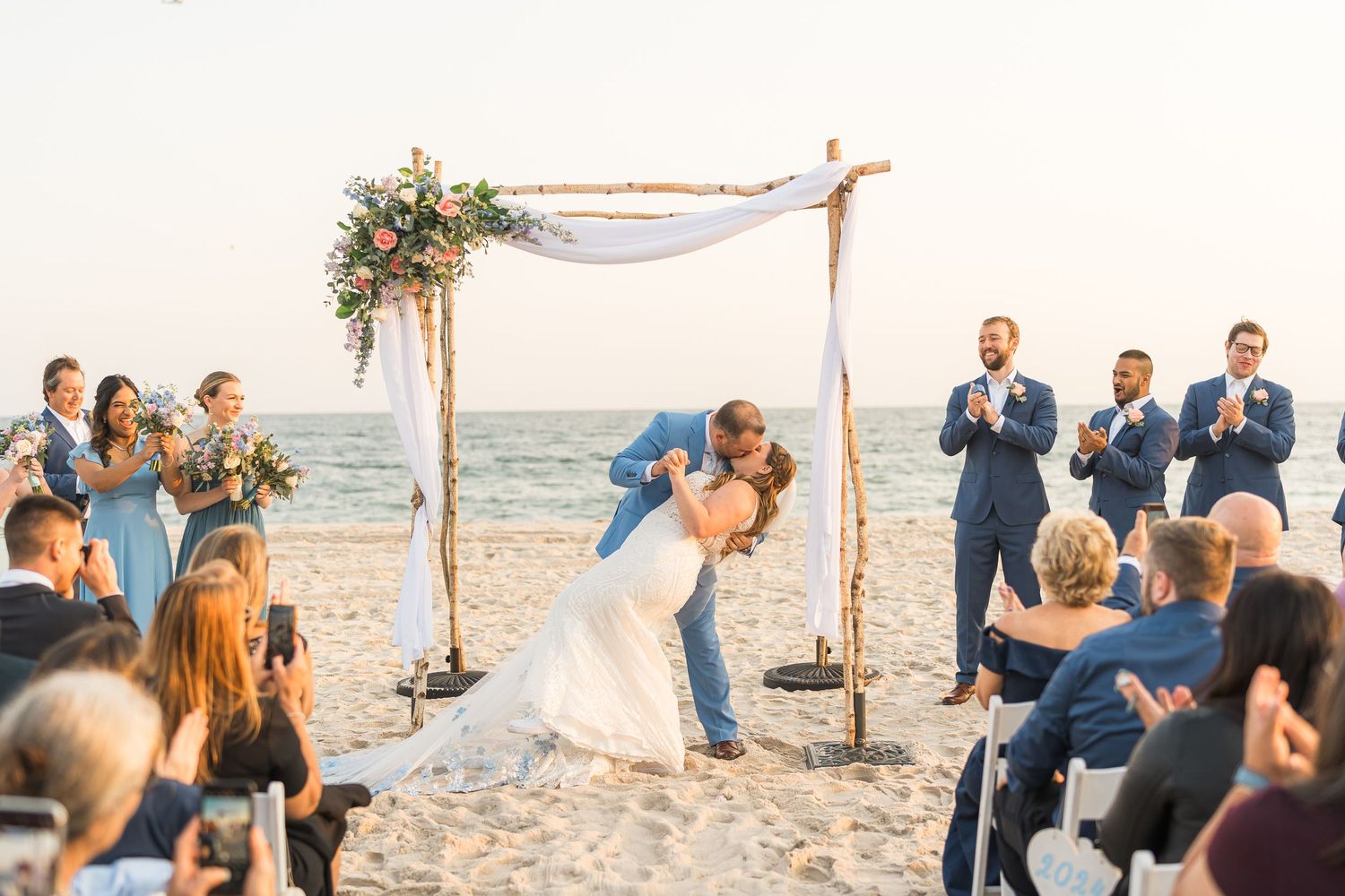 Beach wedding ceremony under wooden arch with blue-clad wedding party and seated guests watching.