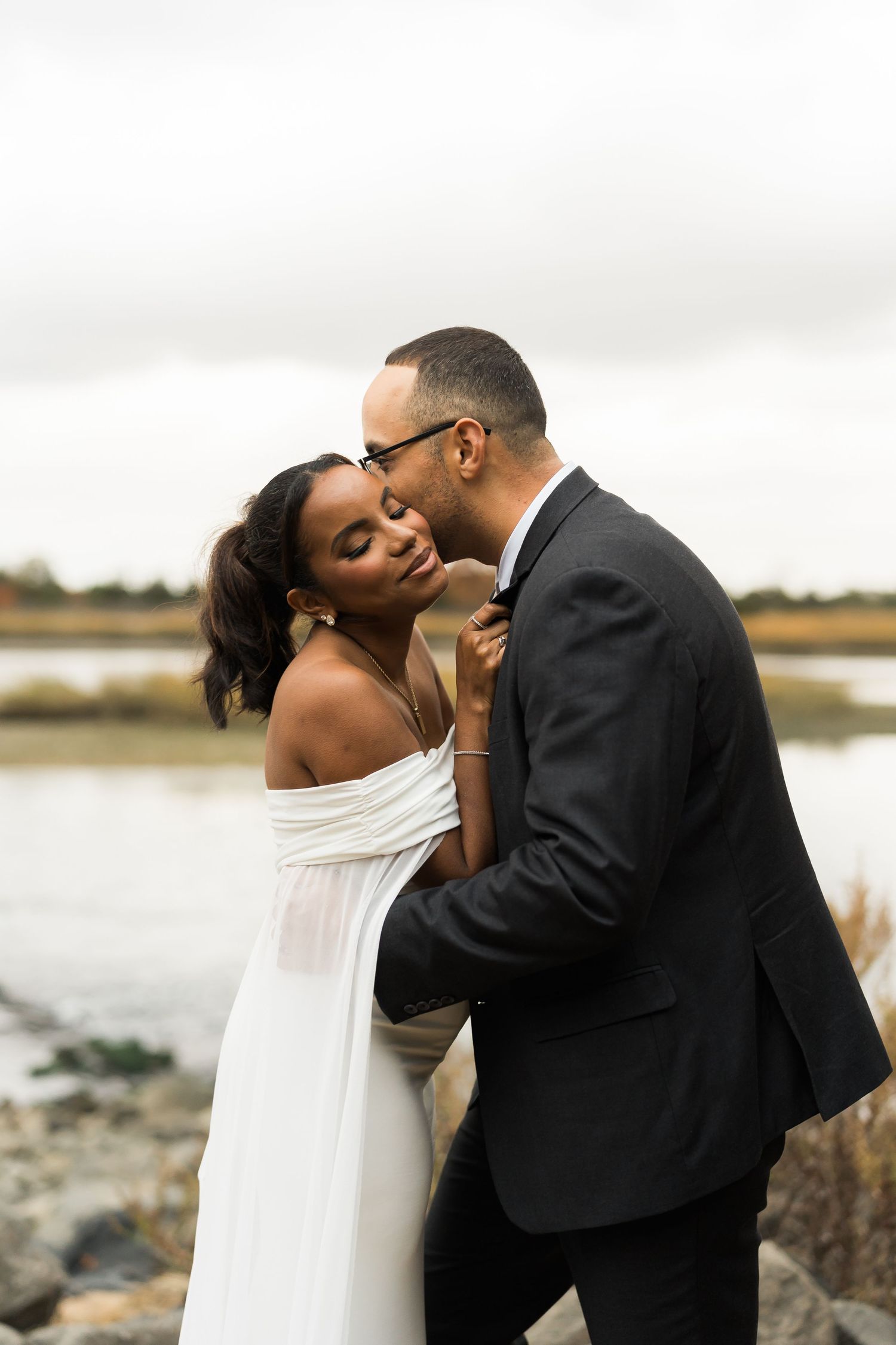 A romantic couple embraces while posing for wedding photos near a scenic waterfront.
