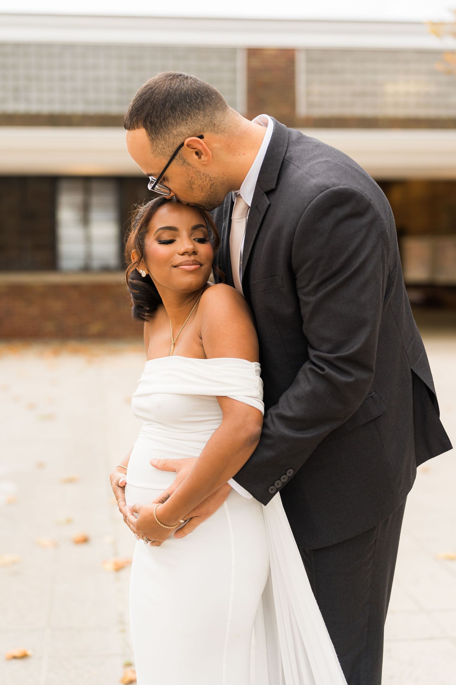 Romantic wedding couple embraces in tender moment outdoors, bride in off-shoulder white gown and groom in classic black suit.