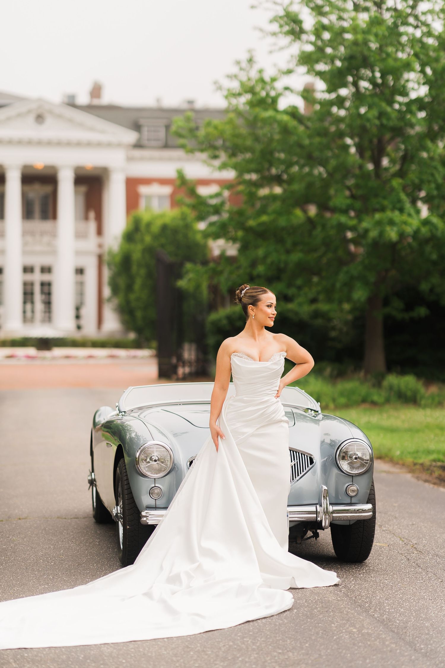 A bride in white gown poses elegantly beside a vintage silver sports car.