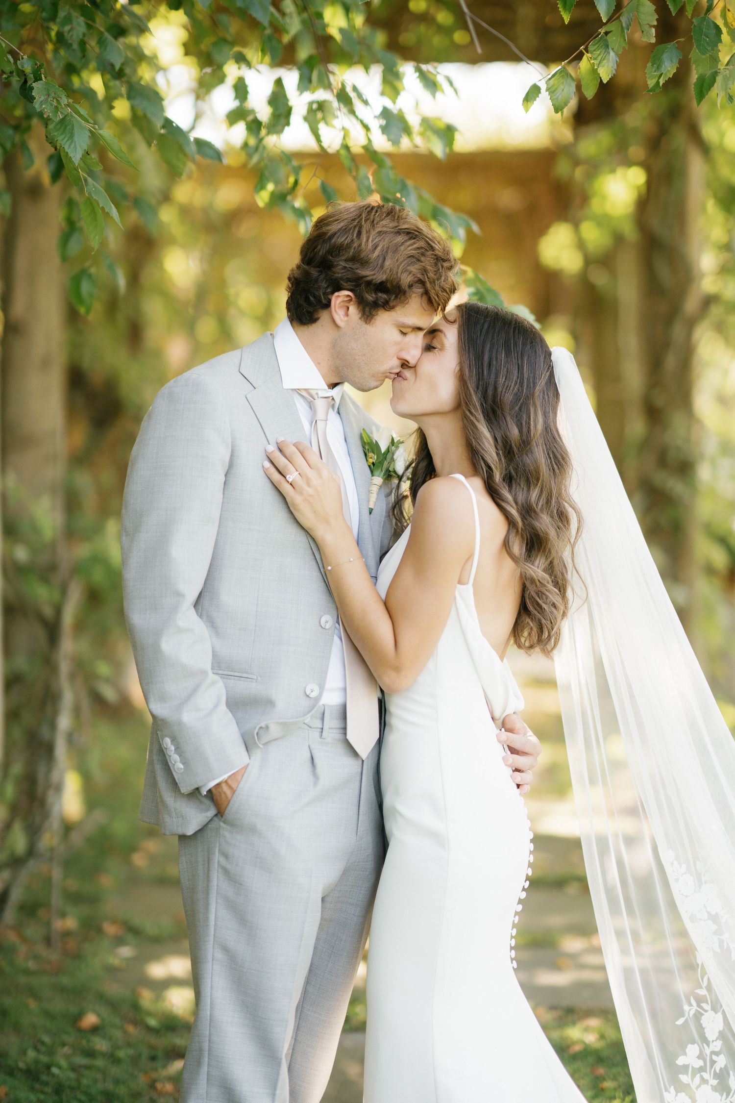 A romantic outdoor wedding photo of a couple sharing a kiss under green trees on a sunny day.