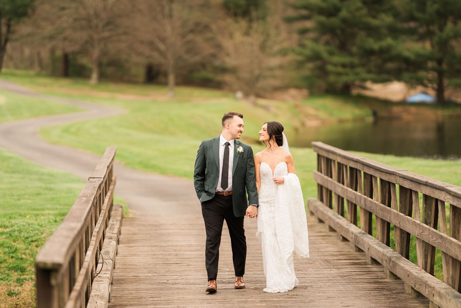 Couple in wedding attire walking together on wooden bridge path through scenic green landscape.