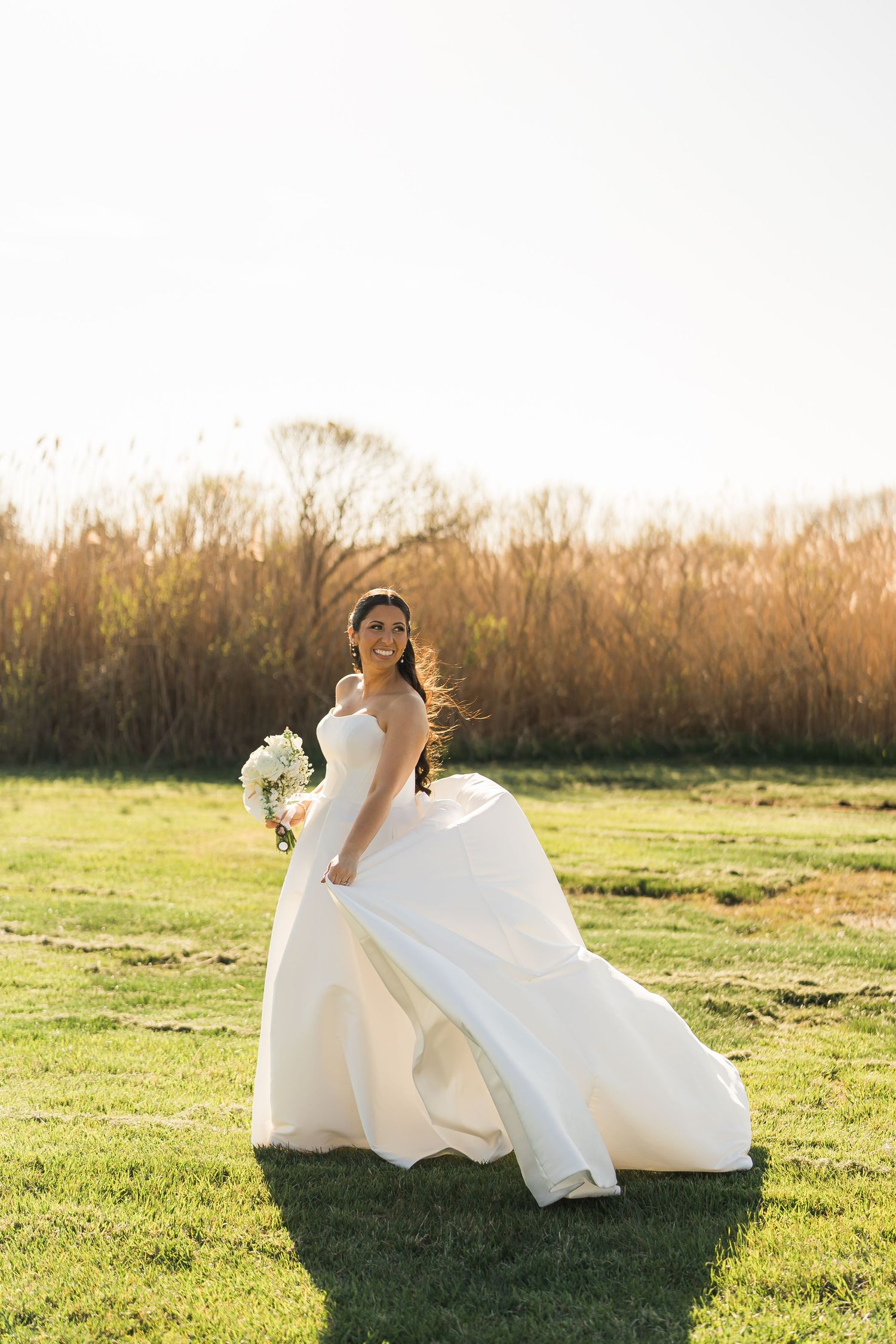 A wedding dress billows in the golden sunlight as its wearer stands in a grassy field at sunset.