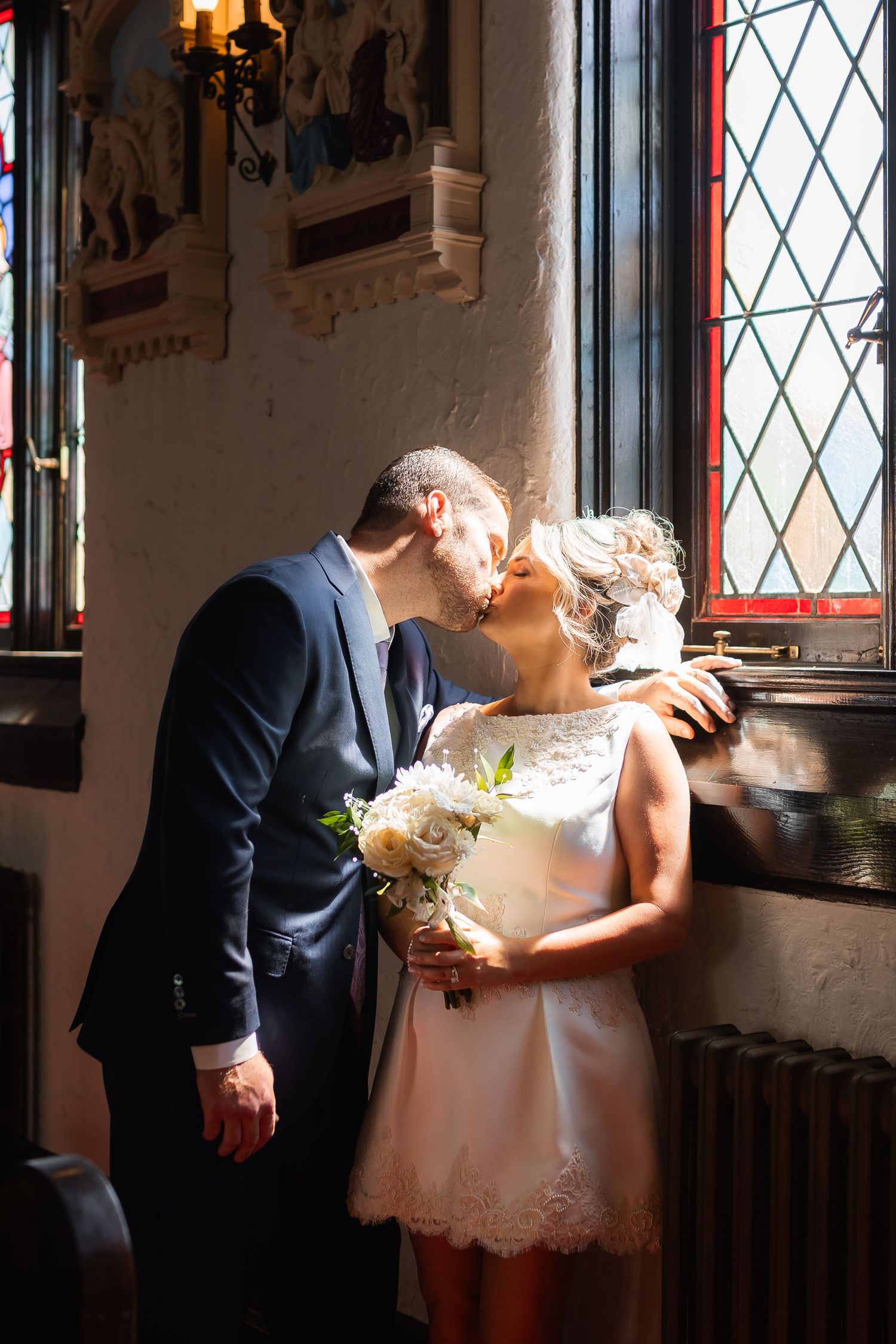 A romantic wedding moment captured by a window with stained glass in an ornate historic building interior.