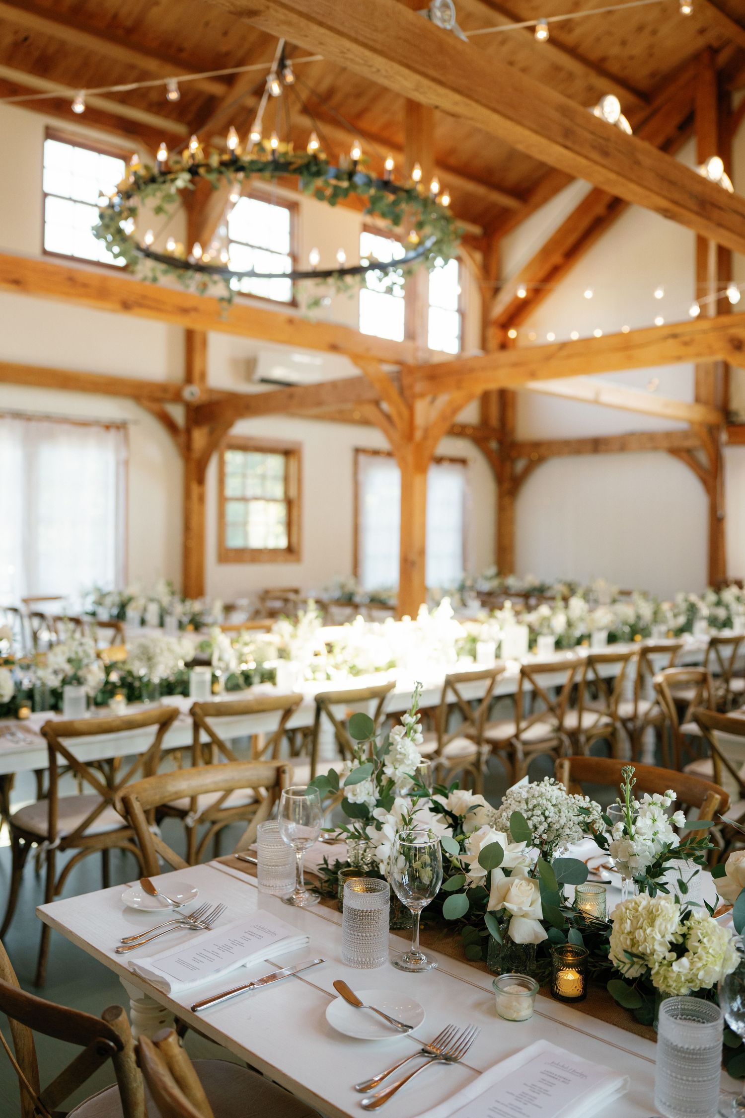 Elegant rustic wedding reception venue with wooden beams, string lights, and long tables decorated with white floral centerpieces.