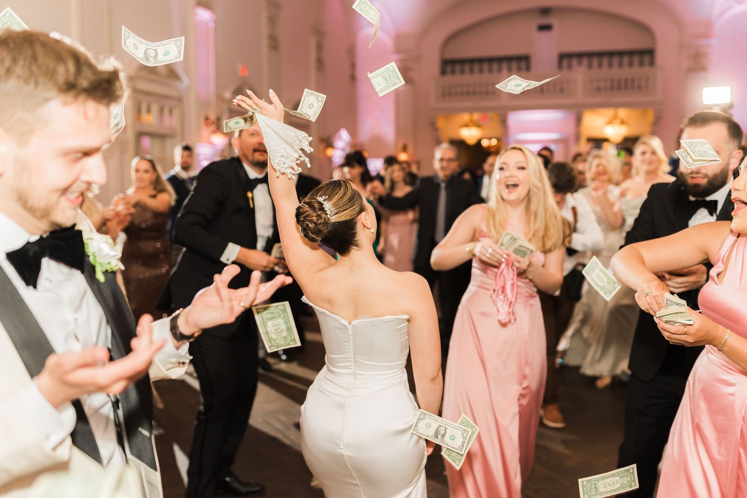 Wedding reception celebration with guests dancing and tossing money in a pink-lit ballroom.