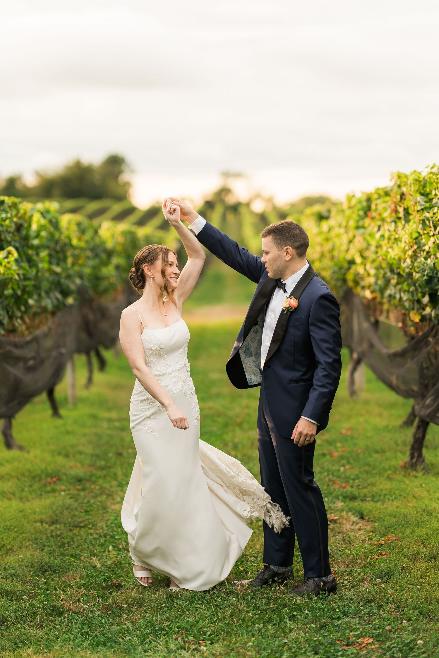 A couple dances together in a vineyard wearing formal wedding attire during golden hour.