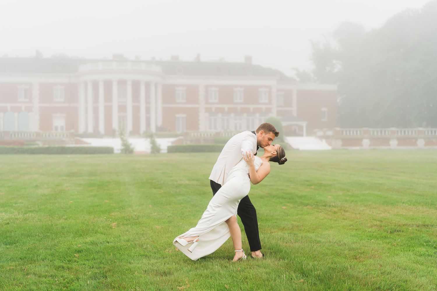 Newlywed couple shares romantic dance pose on misty lawn with elegant mansion in background.