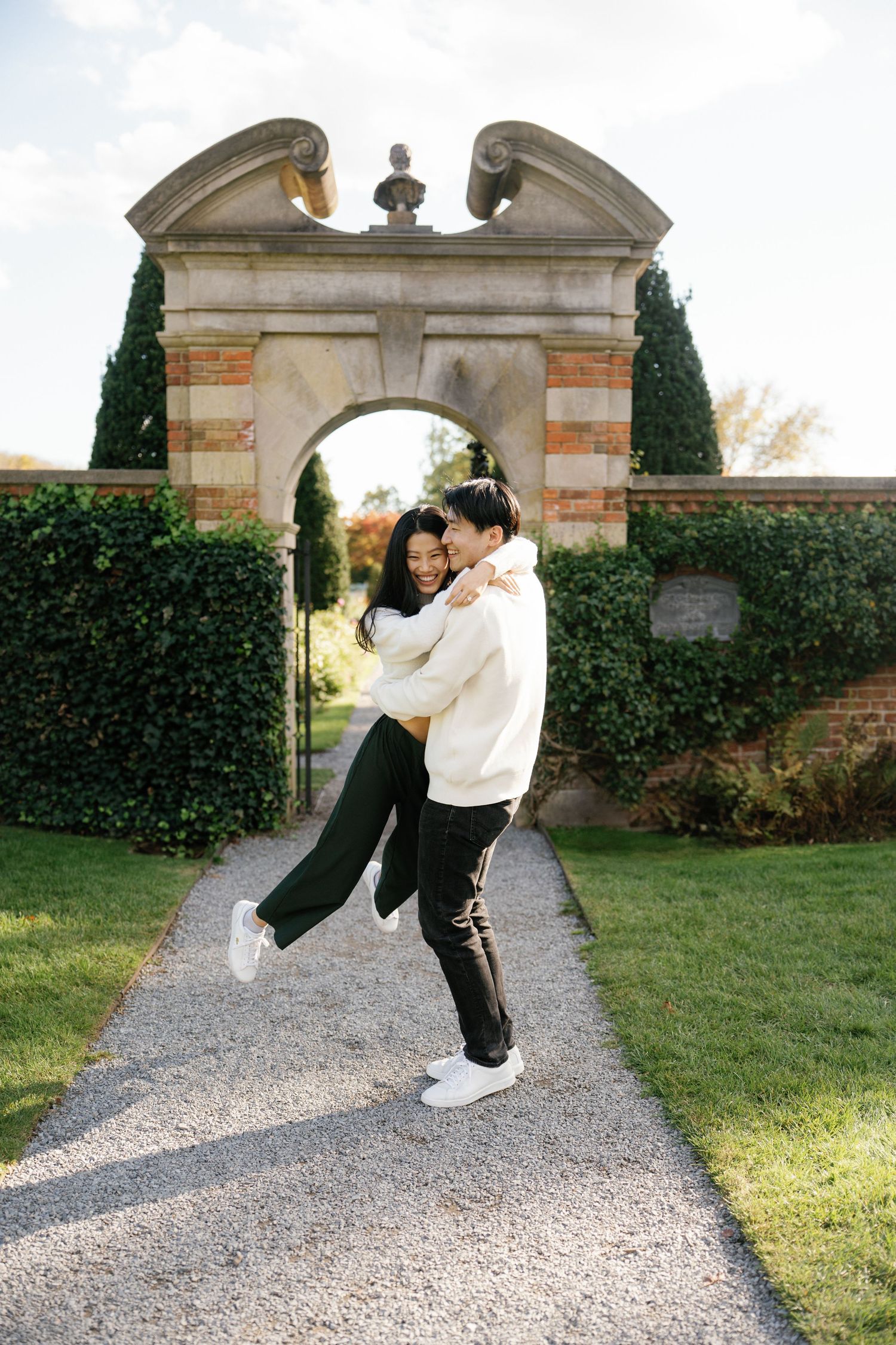 A couple in black and white outfits dance playfully on a gravel path in front of an elegant stone archway.
