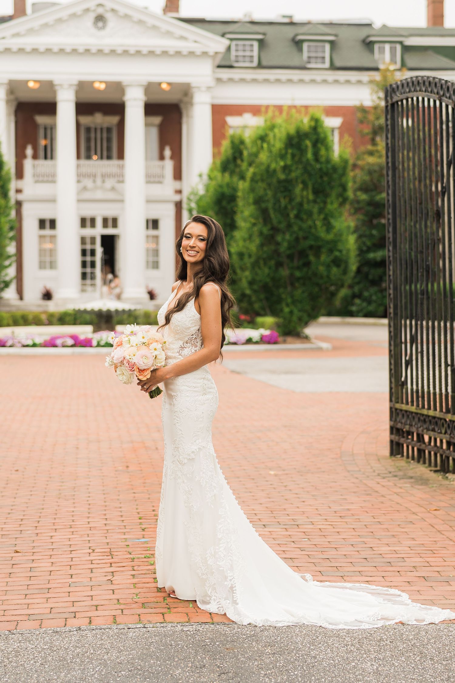Beautiful bride in fitted lace wedding gown poses with peach bouquet in front of elegant mansion with white columns.