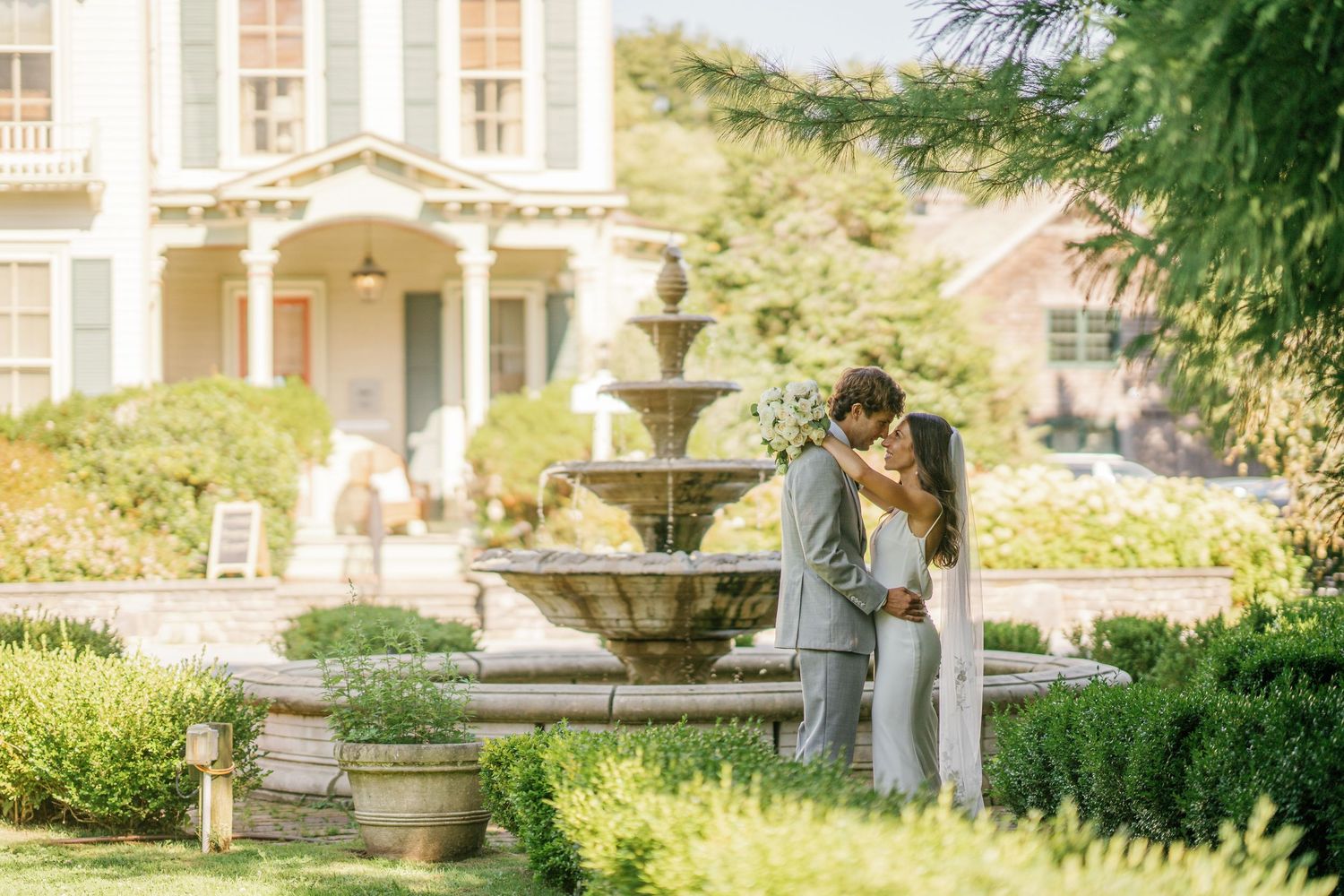 A couple sits together on a stone bench in a lush garden setting with ornate architecture in the background.