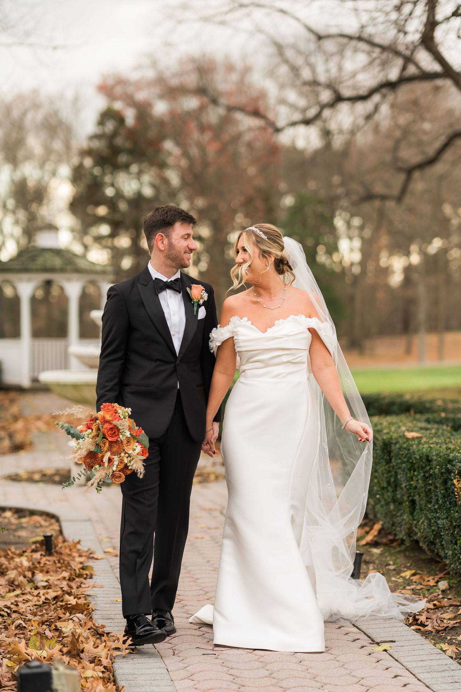 An autumn wedding couple walking together outdoors near a white gazebo surrounded by fallen leaves.