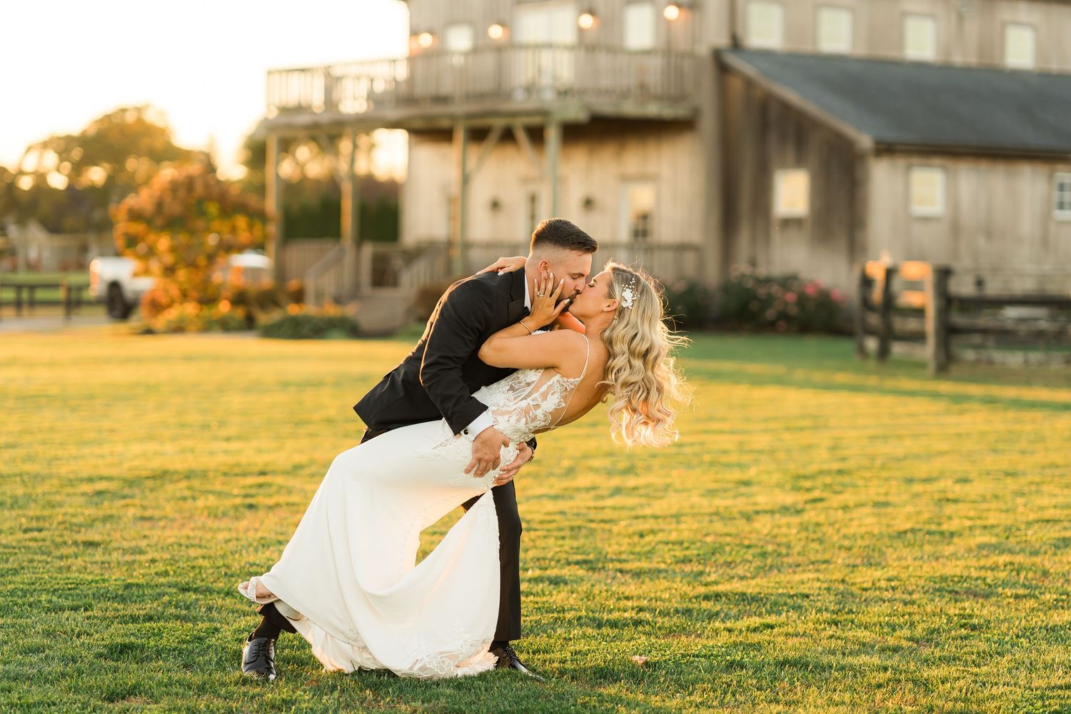 A romantic couple in wedding attire dips dramatically on a sunlit lawn with buildings in the background at golden hour.