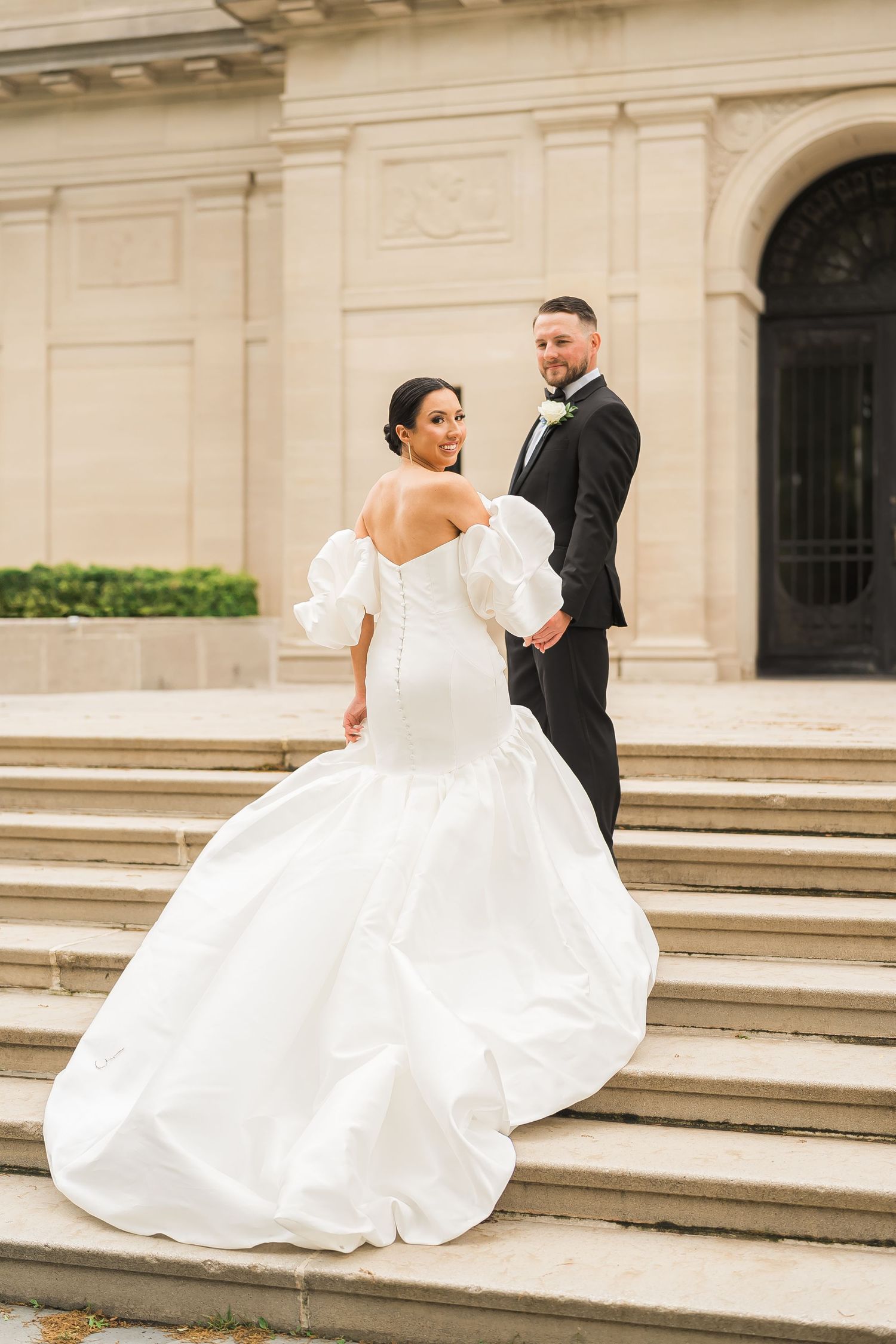 A couple poses on grand stone steps with a flowing white wedding gown.