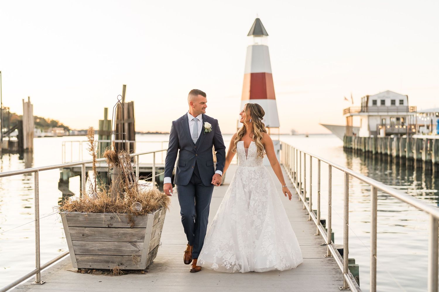 A couple walks along a marina boardwalk with a lighthouse in the background.