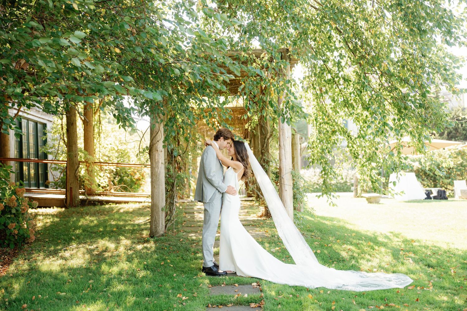 A couple shares a romantic moment under lush green trees in a garden setting with a flowing white wedding veil.