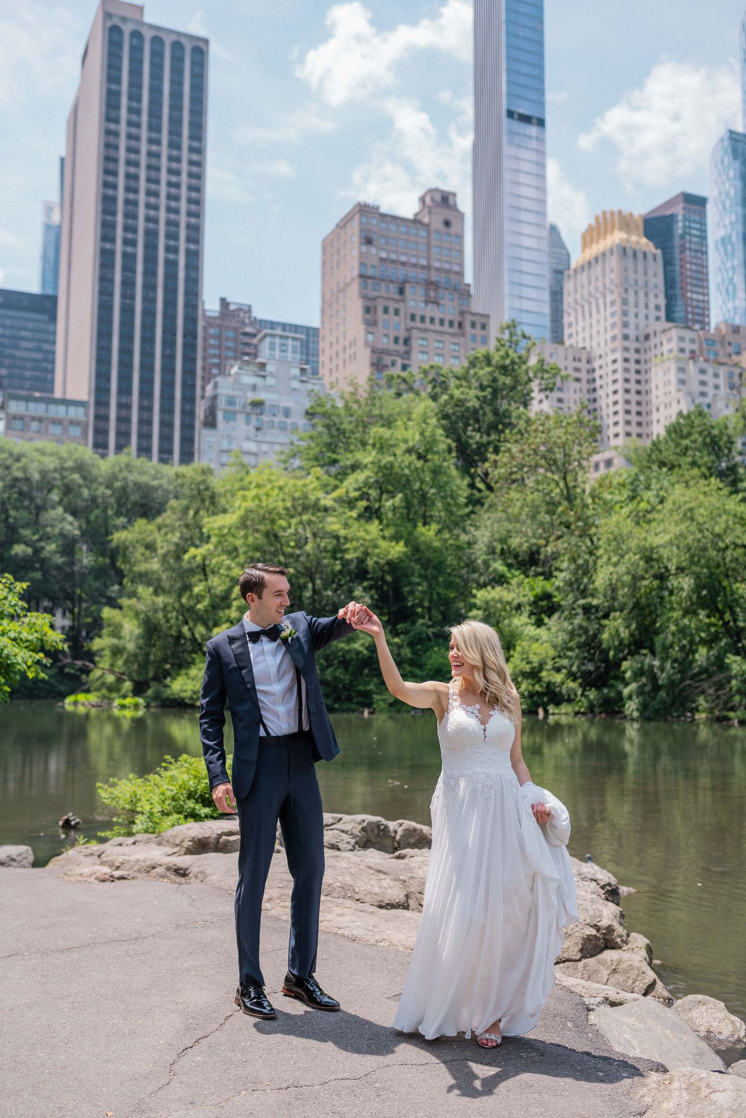 Wedding couple poses playfully near a lake in Central Park with Manhattan skyline in background.