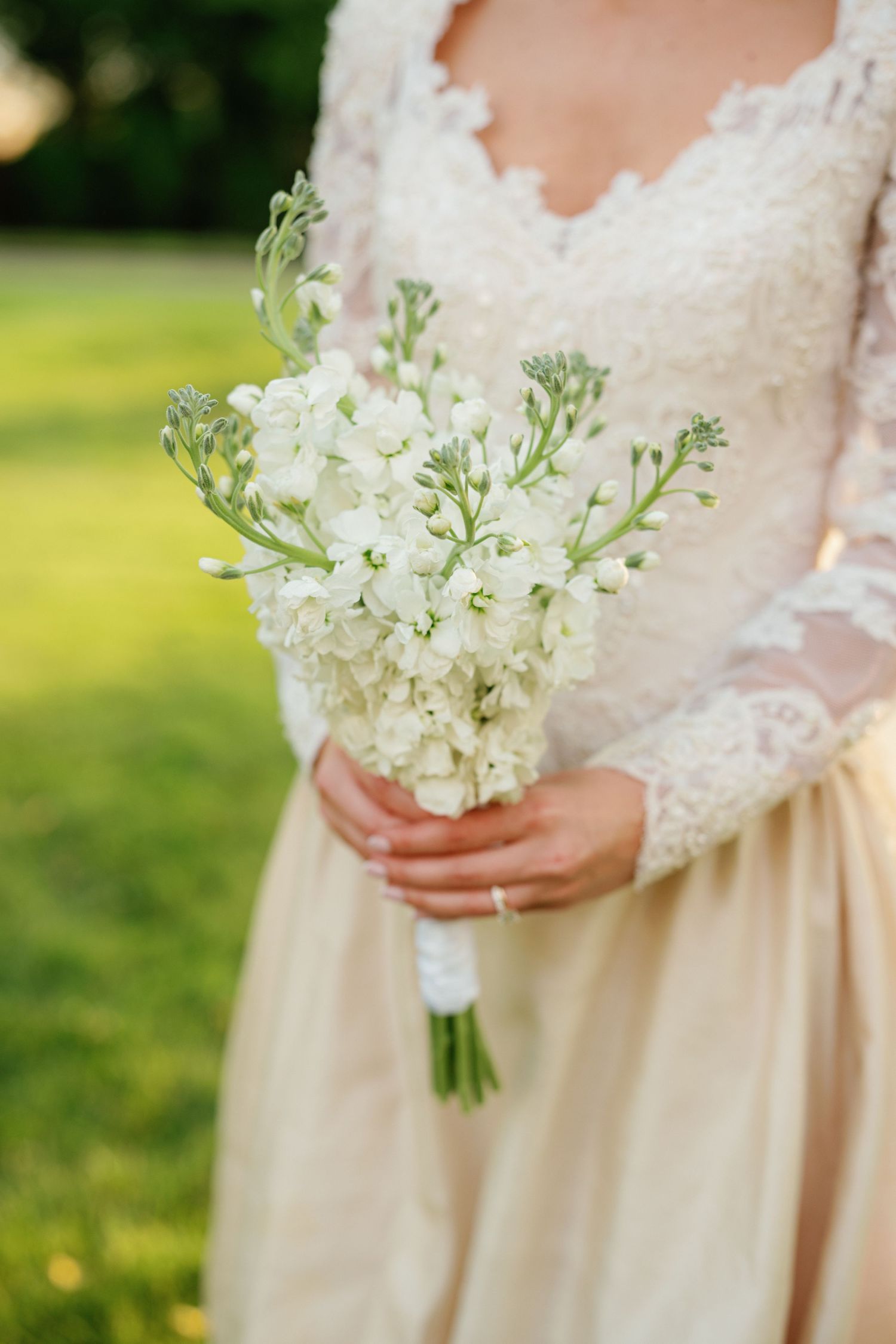 A delicate white floral bouquet held against a cream-colored lace wedding dress in an outdoor garden setting.