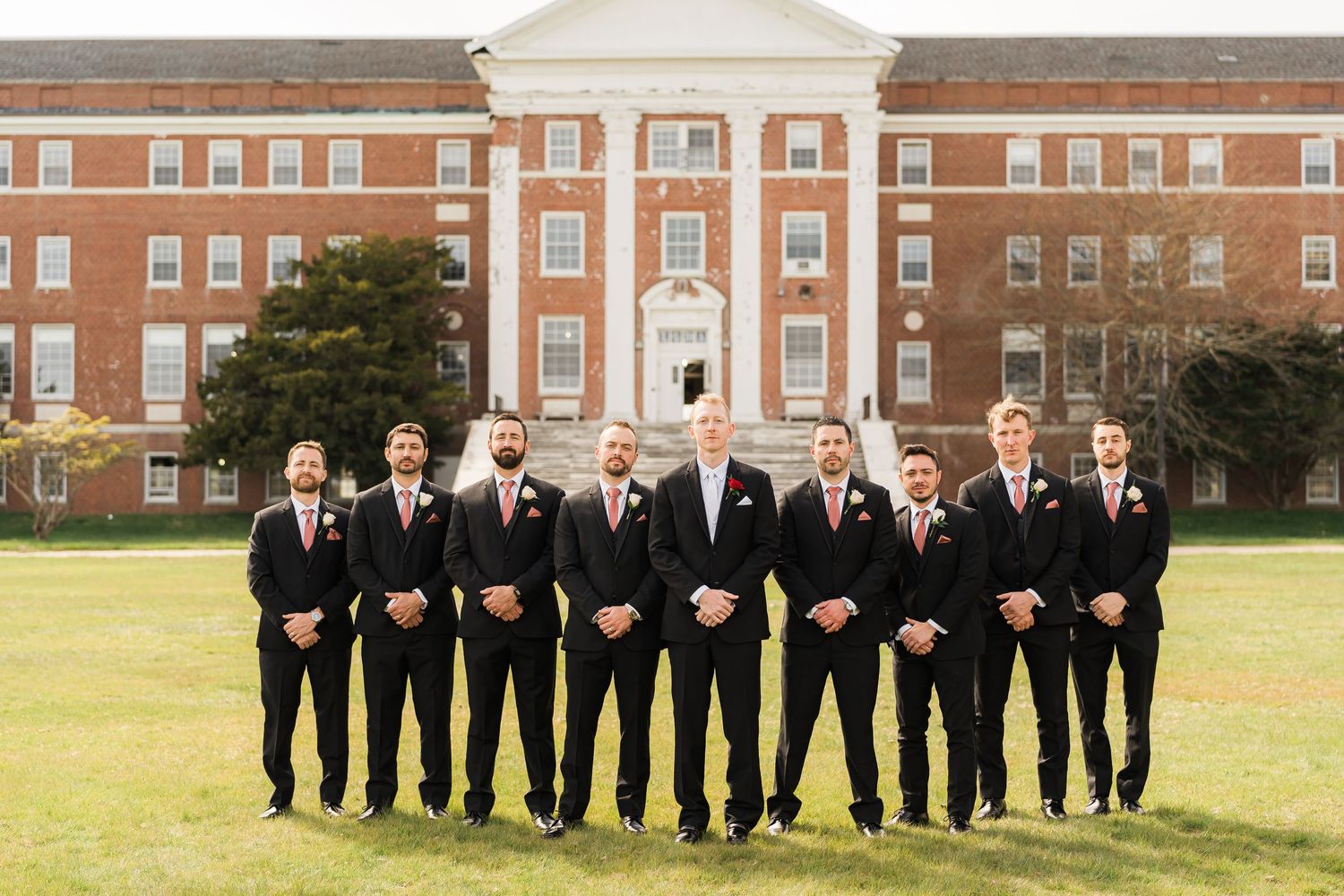 Groomsmen in matching black suits and ties pose together on lawn in front of brick building.