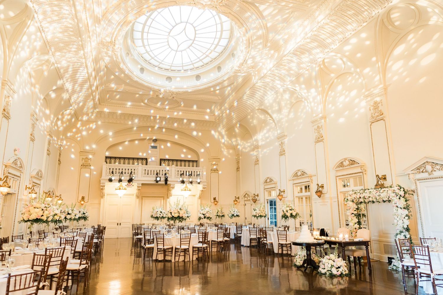 An elegant ballroom features ornate ceiling details and is set up for a wedding reception with round tables.