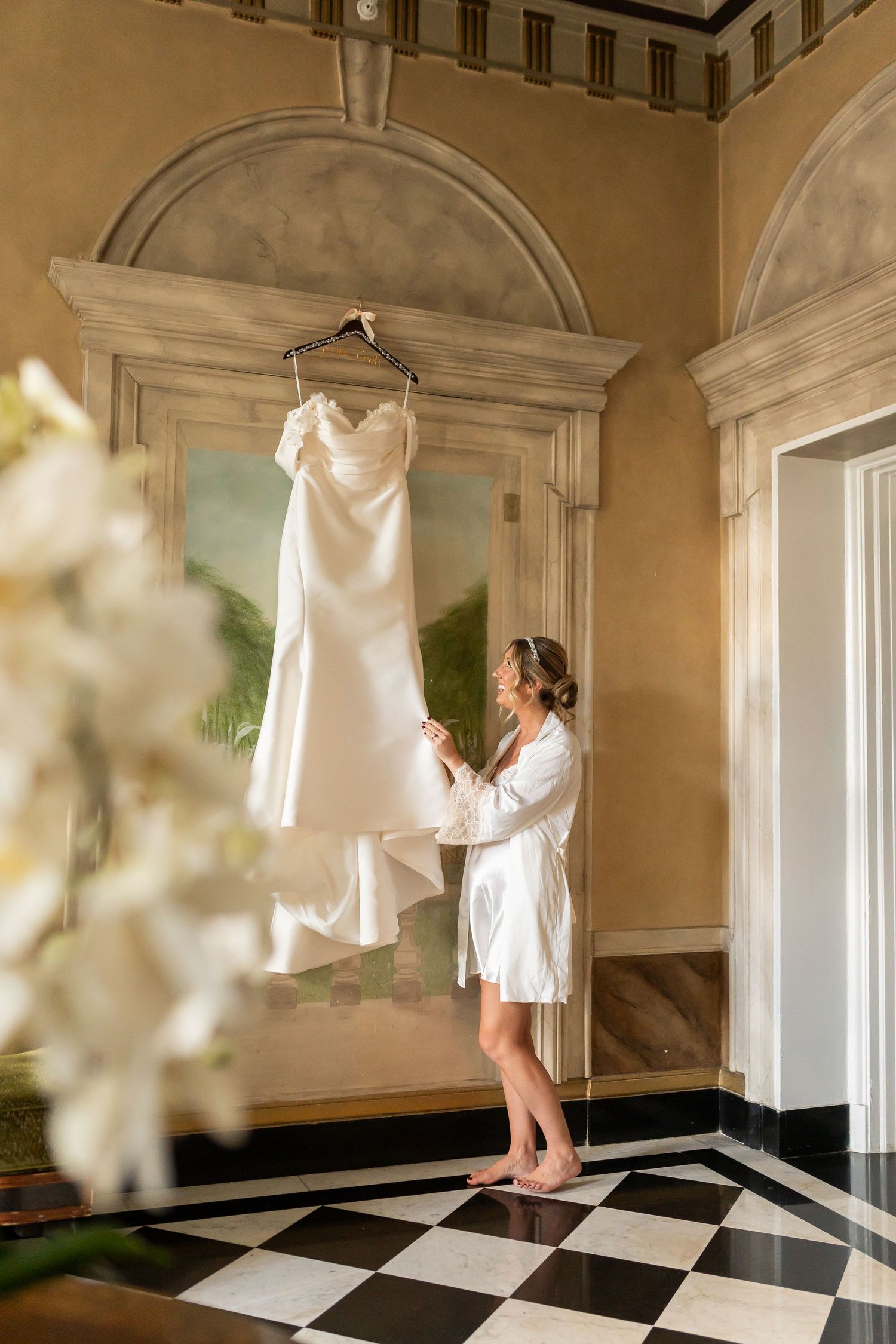 A white wedding dress hangs elegantly in an ornate room with a black and white checkered floor.