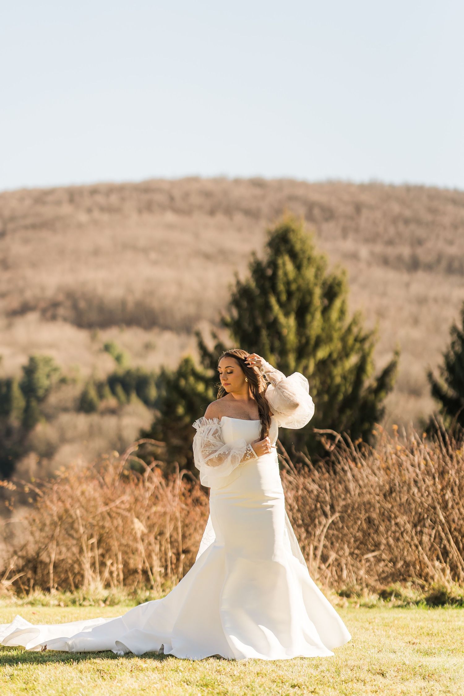 A bride in a long-sleeved white wedding gown poses outdoors against a scenic hillside with evergreen trees.
