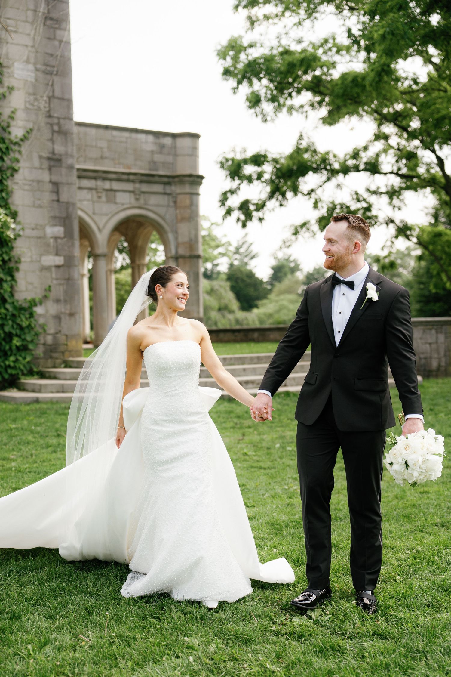 A couple in wedding attire playfully poses on a grassy lawn near stone archways on their special day.