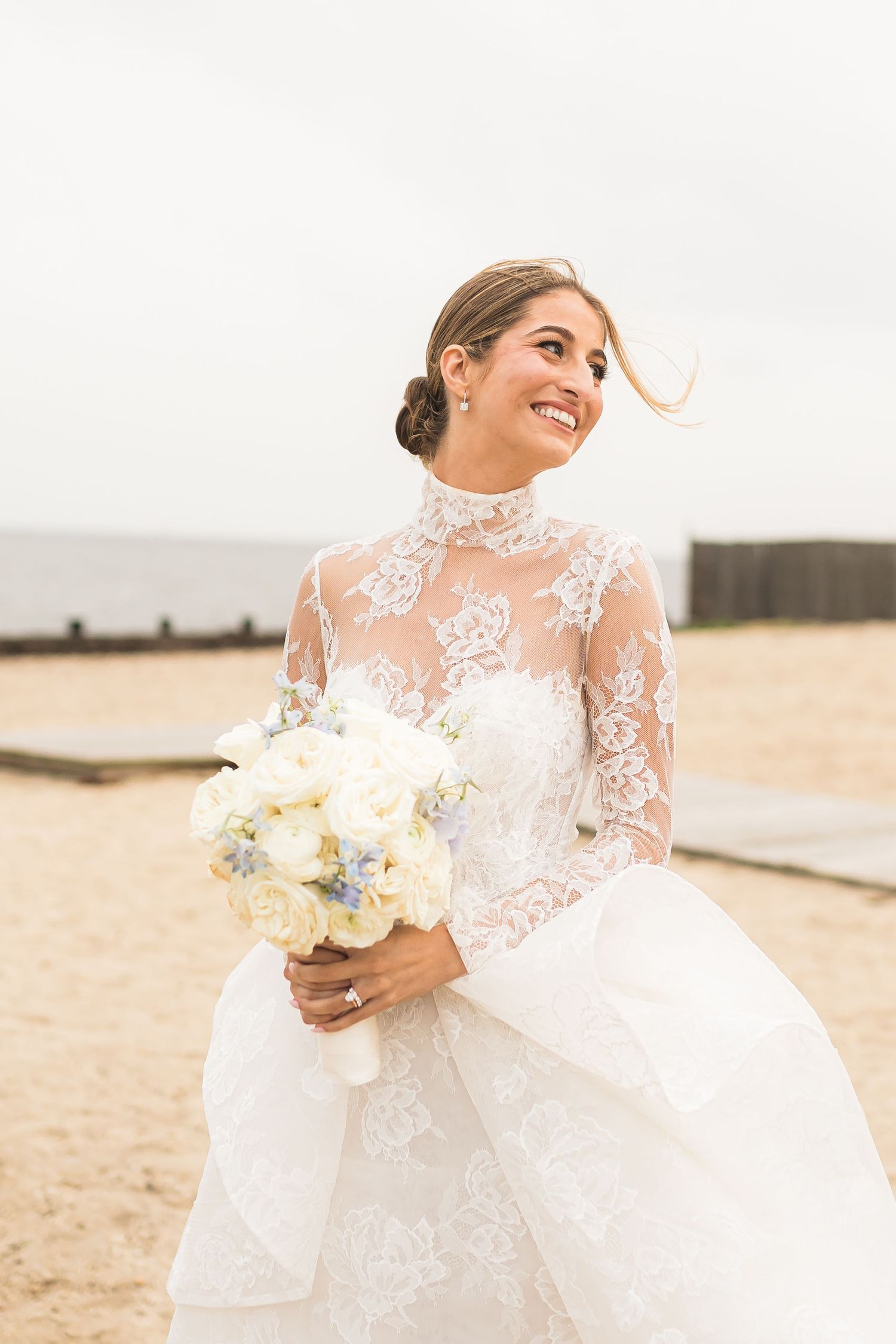 Bride in lace gown holds white bouquet while smiling on beach.