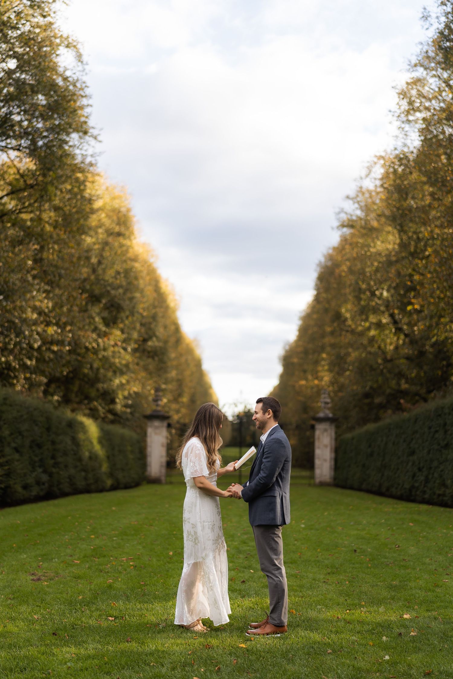 A couple stands together on a manicured lawn path lined with tall autumn trees and hedges creating a garden tunnel effect.