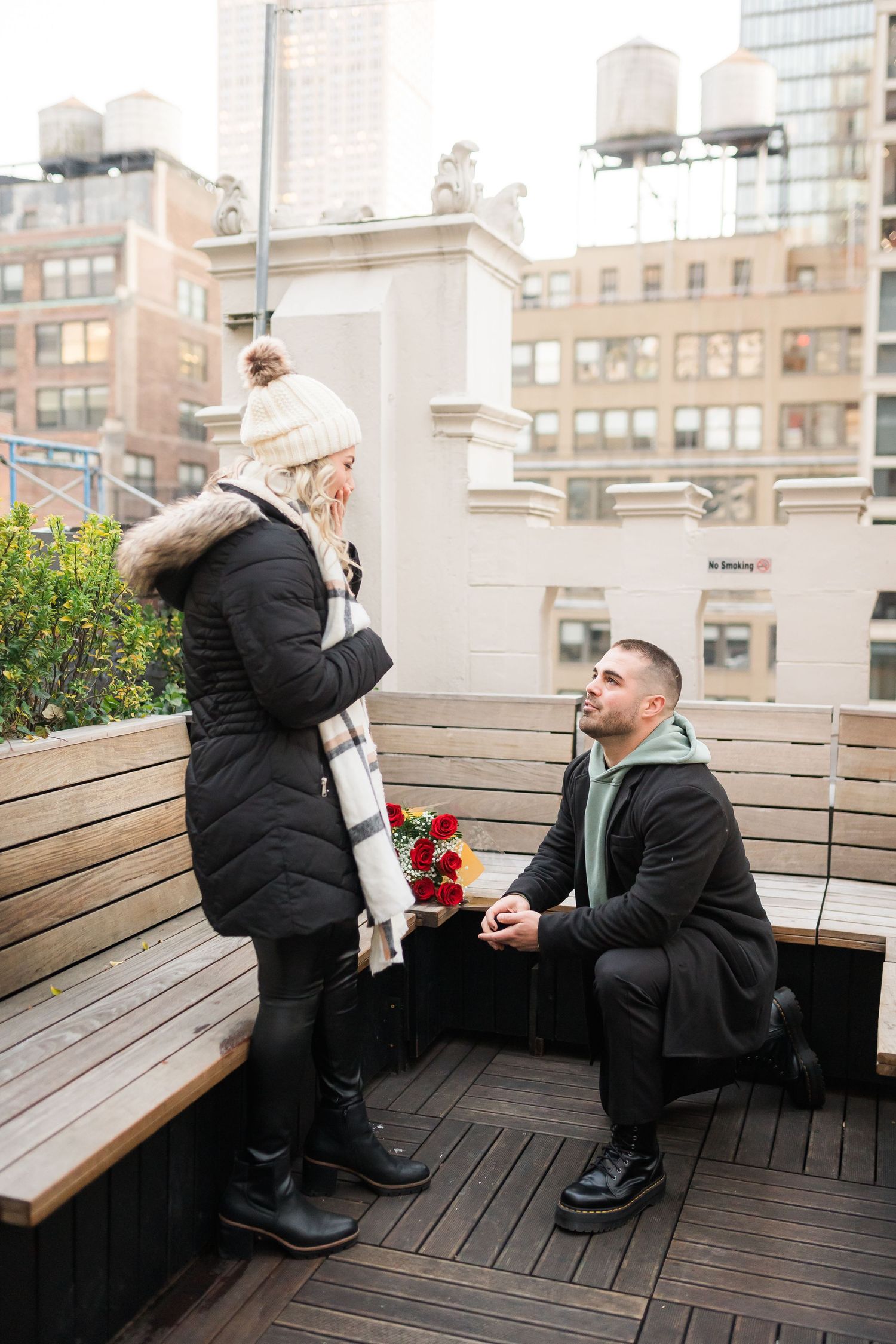 A romantic rooftop proposal moment with city buildings in the background and red roses.