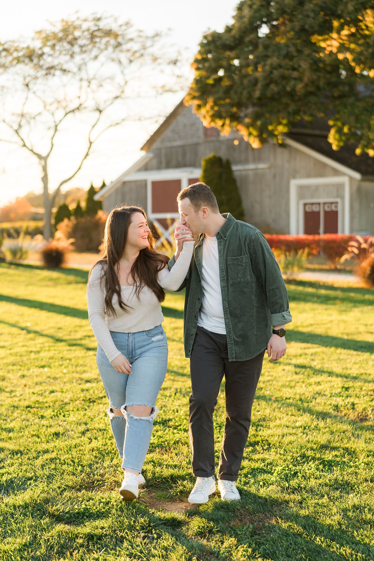 Couple laughing together on grassy lawn with rustic barn in golden afternoon sunlight.