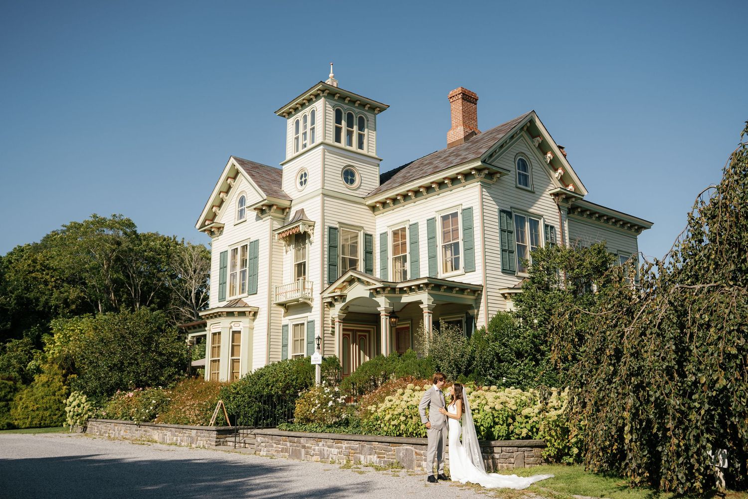 Elegant white Victorian mansion with wraparound porch and manicured gardens photographed at sunset.