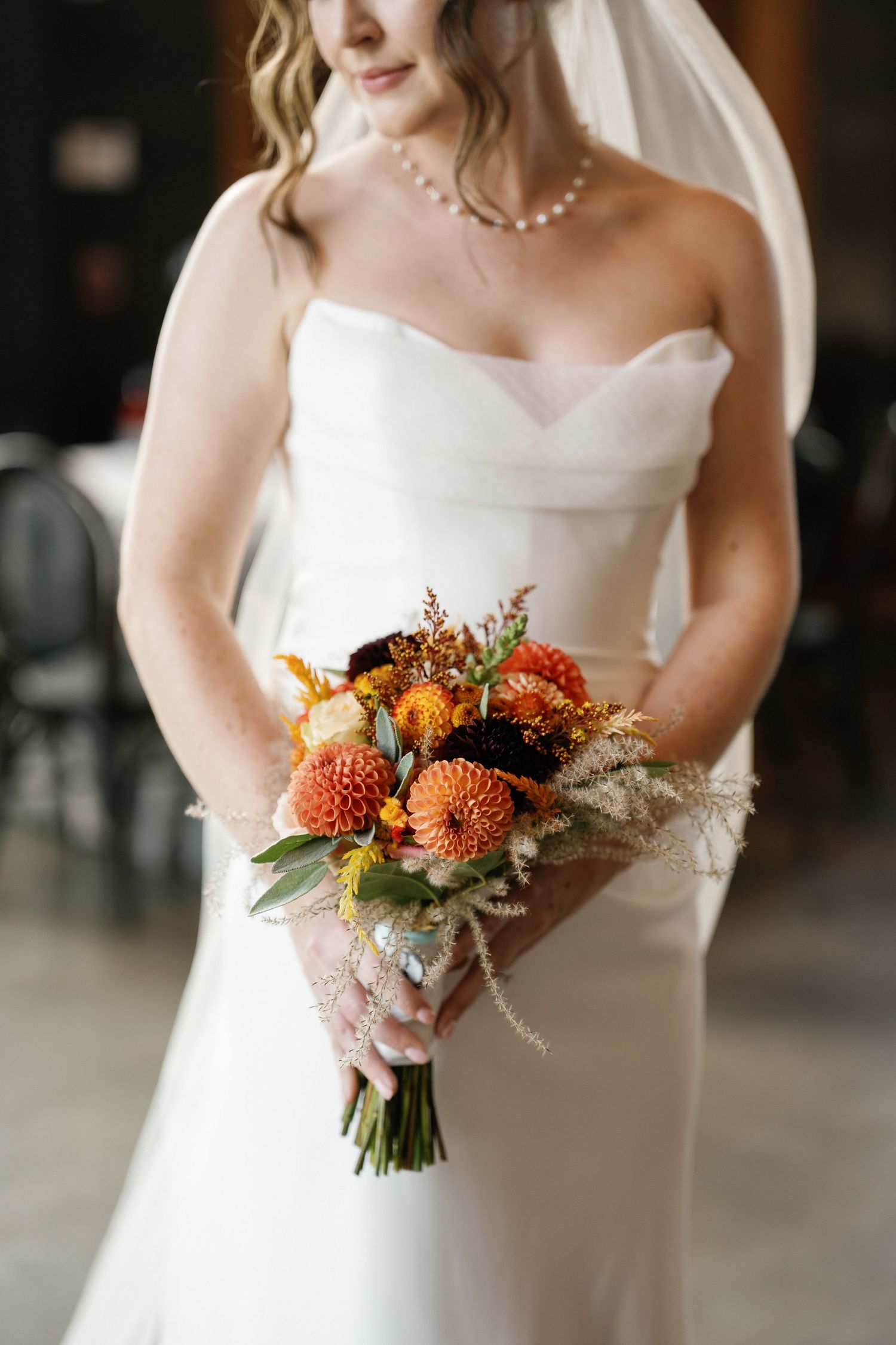 A bride in a white strapless wedding gown holds a vibrant orange and coral fall wedding bouquet against a dark background.