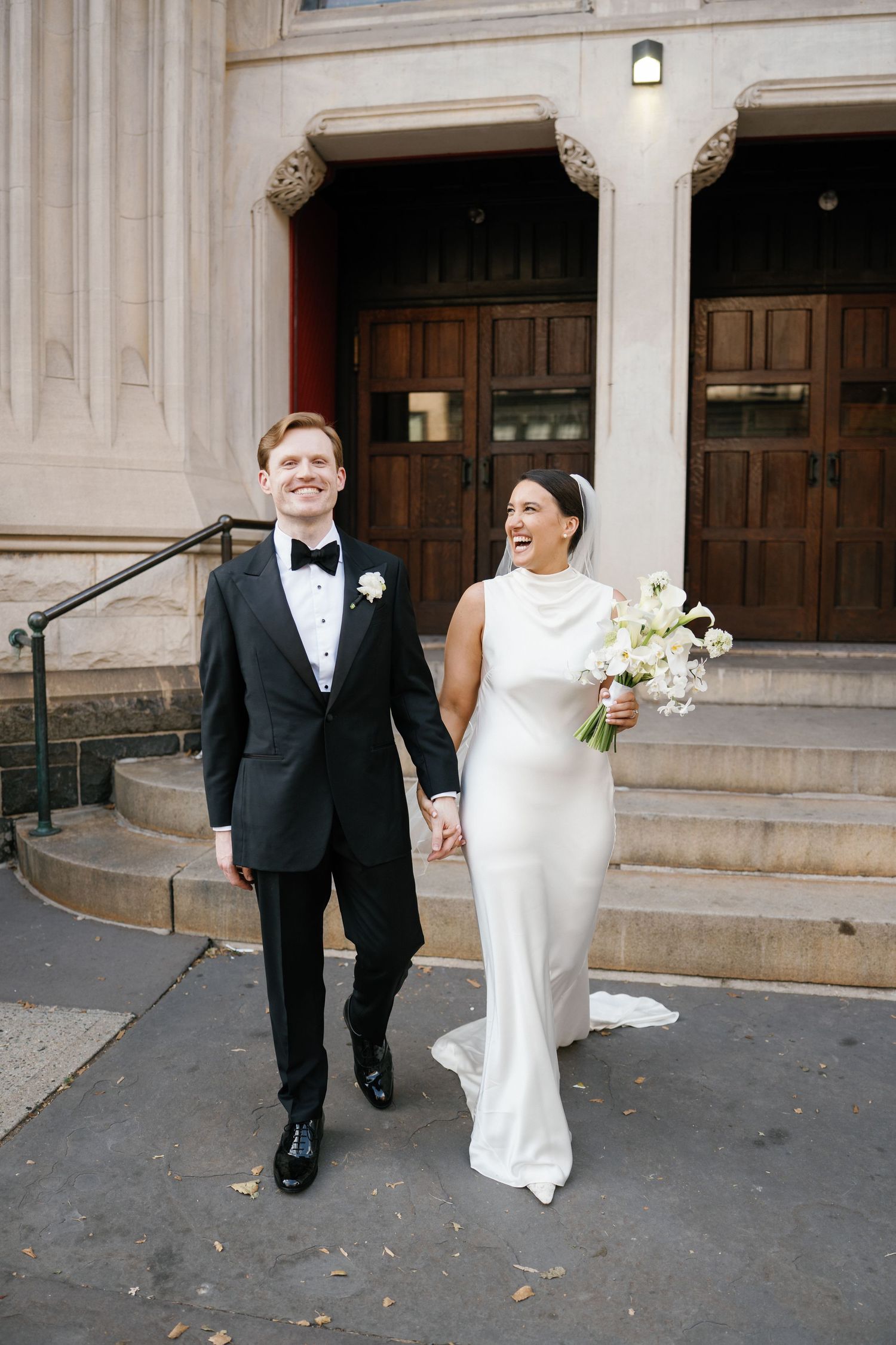 A couple in formal wedding attire laughs and walks together outside a grand building with stone columns.