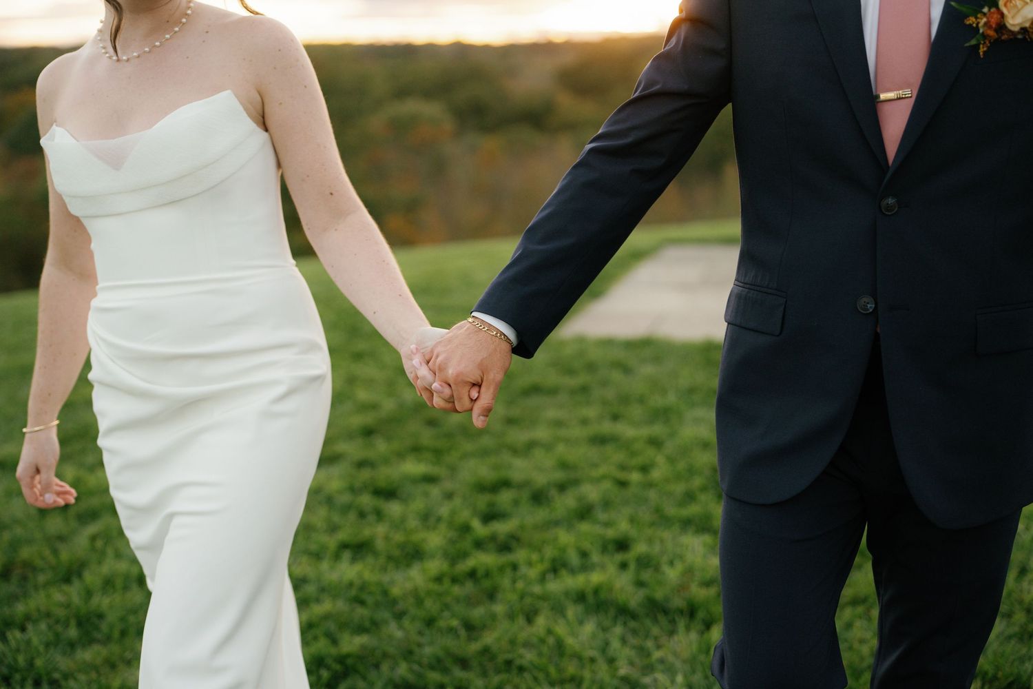Newlywed couple holding hands while walking on lush green grass during sunset at their outdoor wedding celebration.
