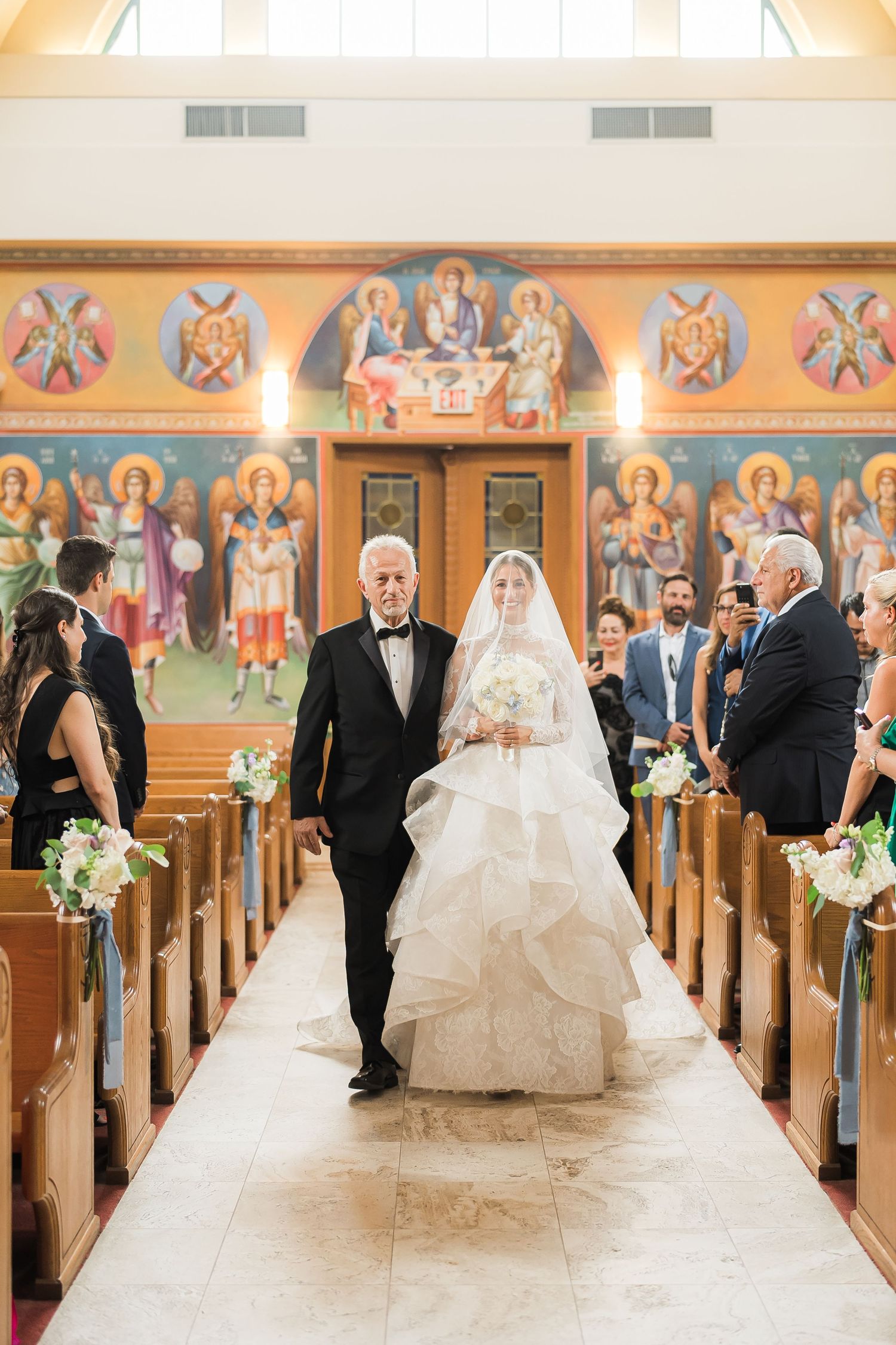 A wedding ceremony in a church with ornate religious murals lining the walls above wooden pews.