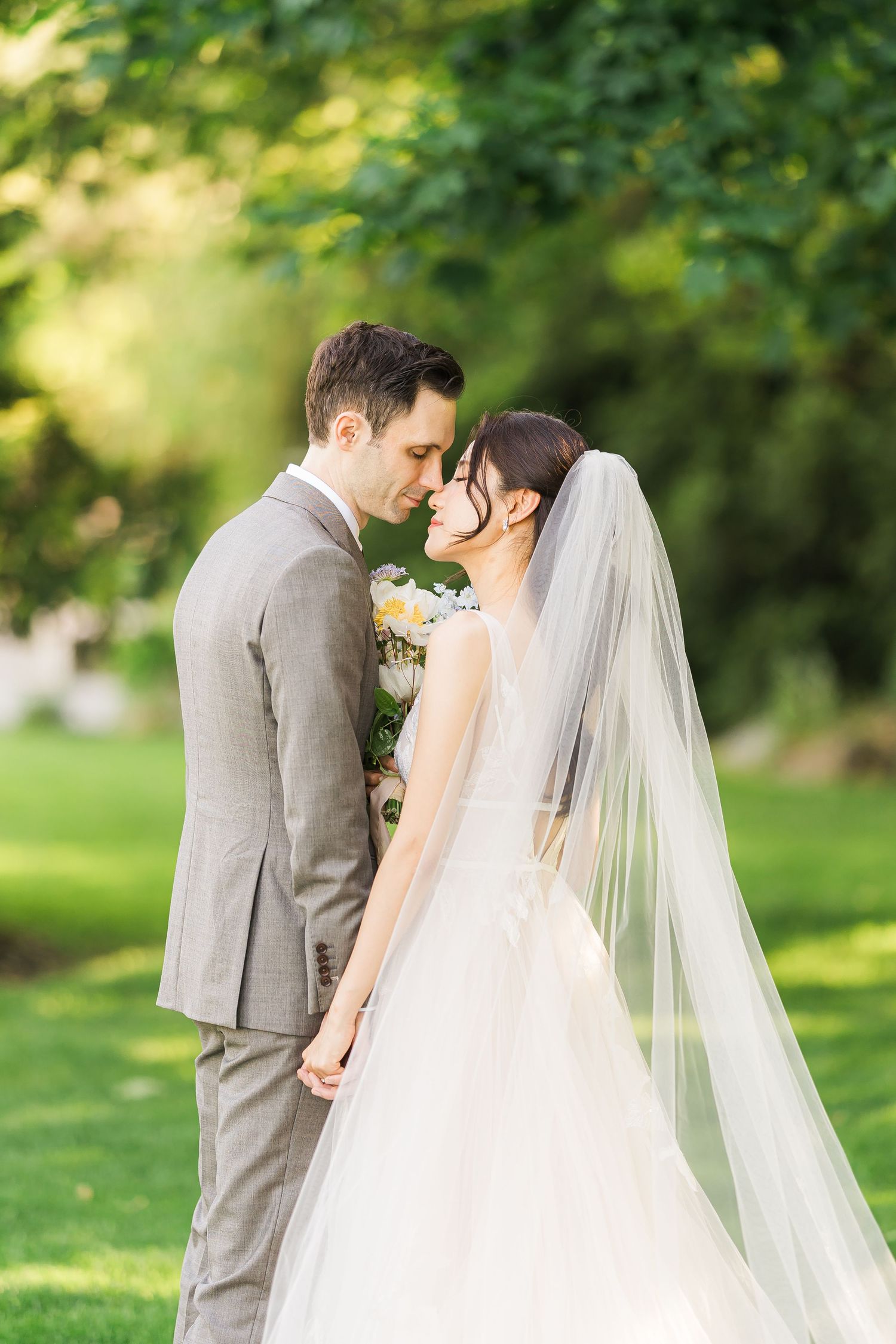 Bride in flowing white dress and veil embraces groom in grey suit during romantic garden wedding photo shoot.