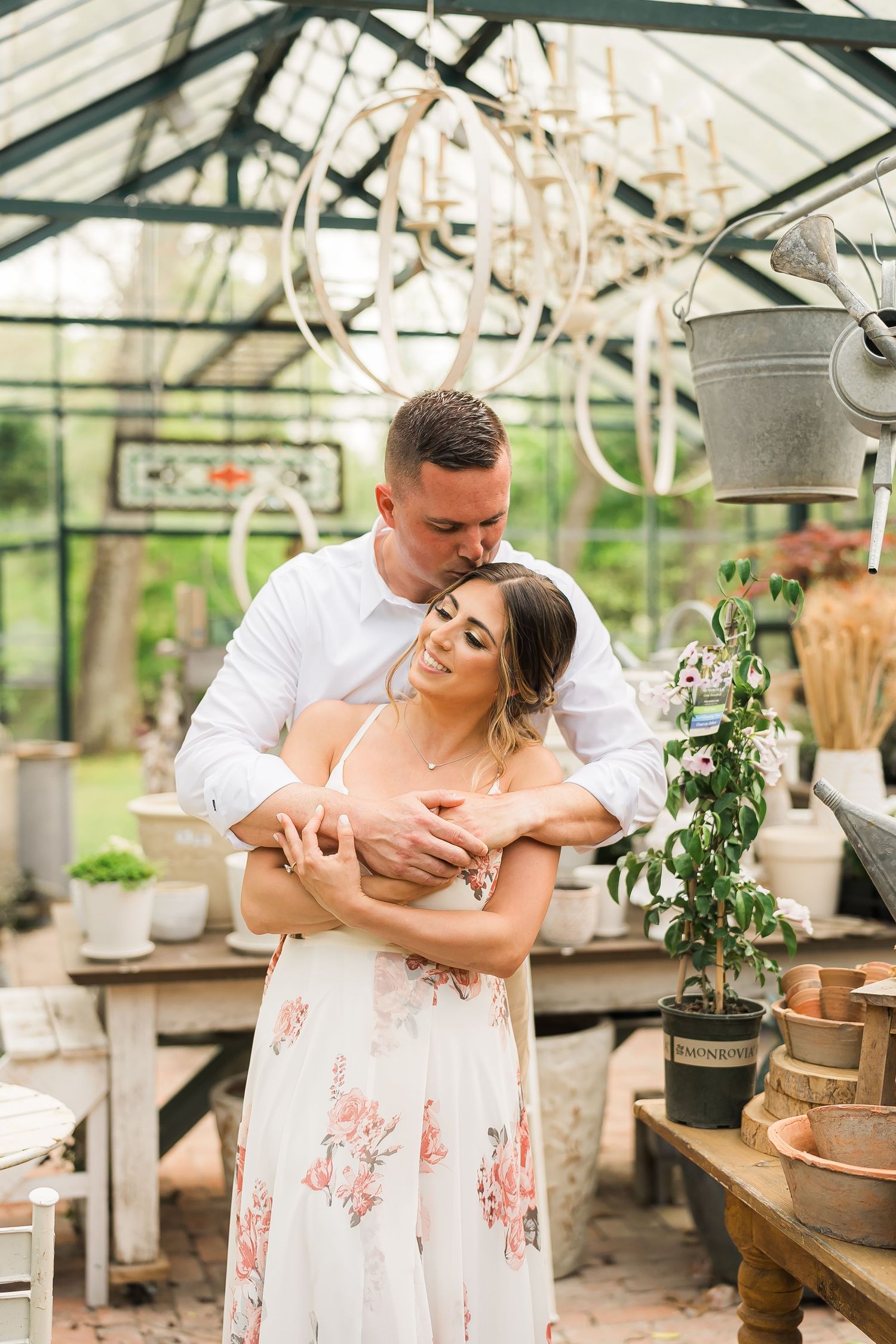 A couple sharing an intimate moment in a sunlit greenhouse filled with plants and hanging lights.