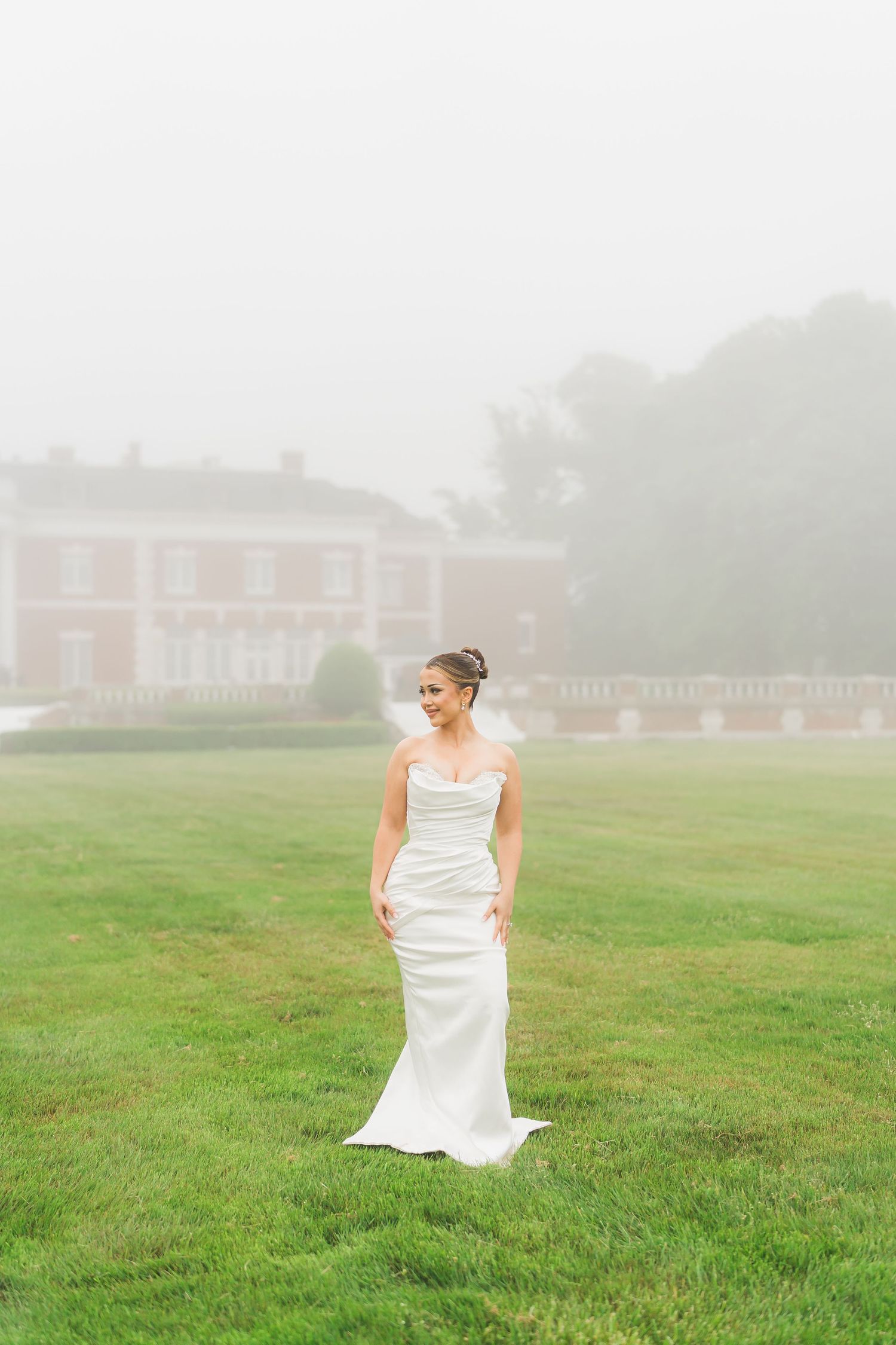 A bride in a fitted white gown stands alone on misty grounds with a mansion in the background.