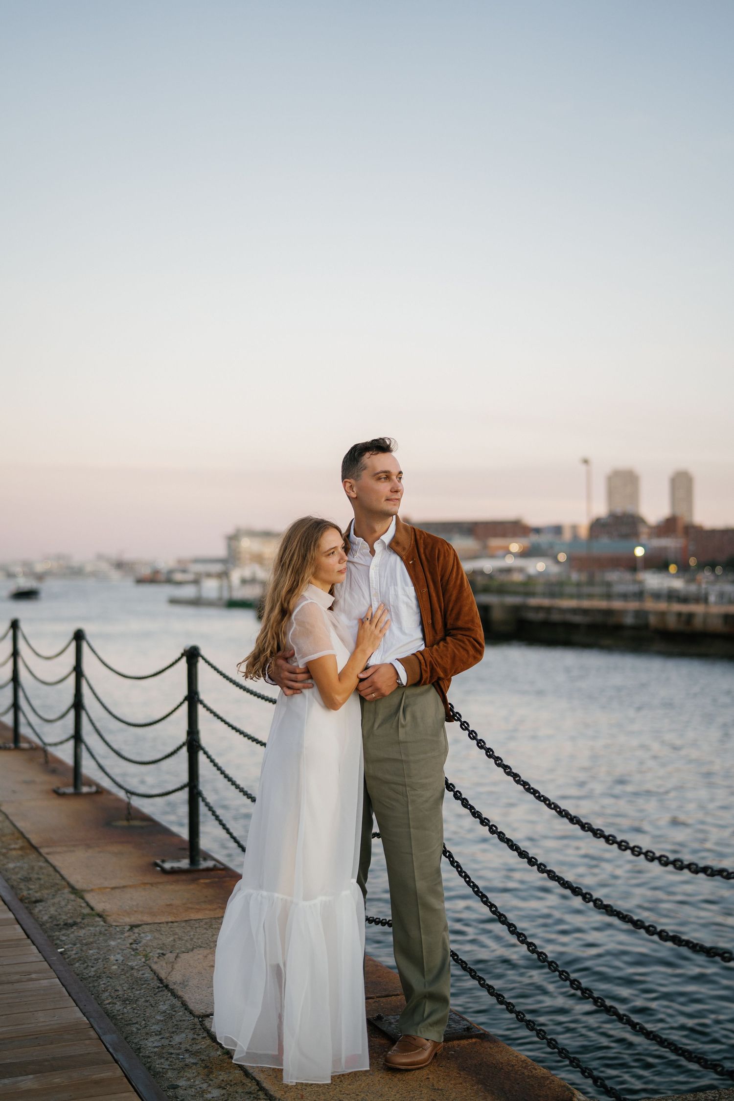 A couple stands together on a waterfront pier at sunset with boats and city buildings in the background.