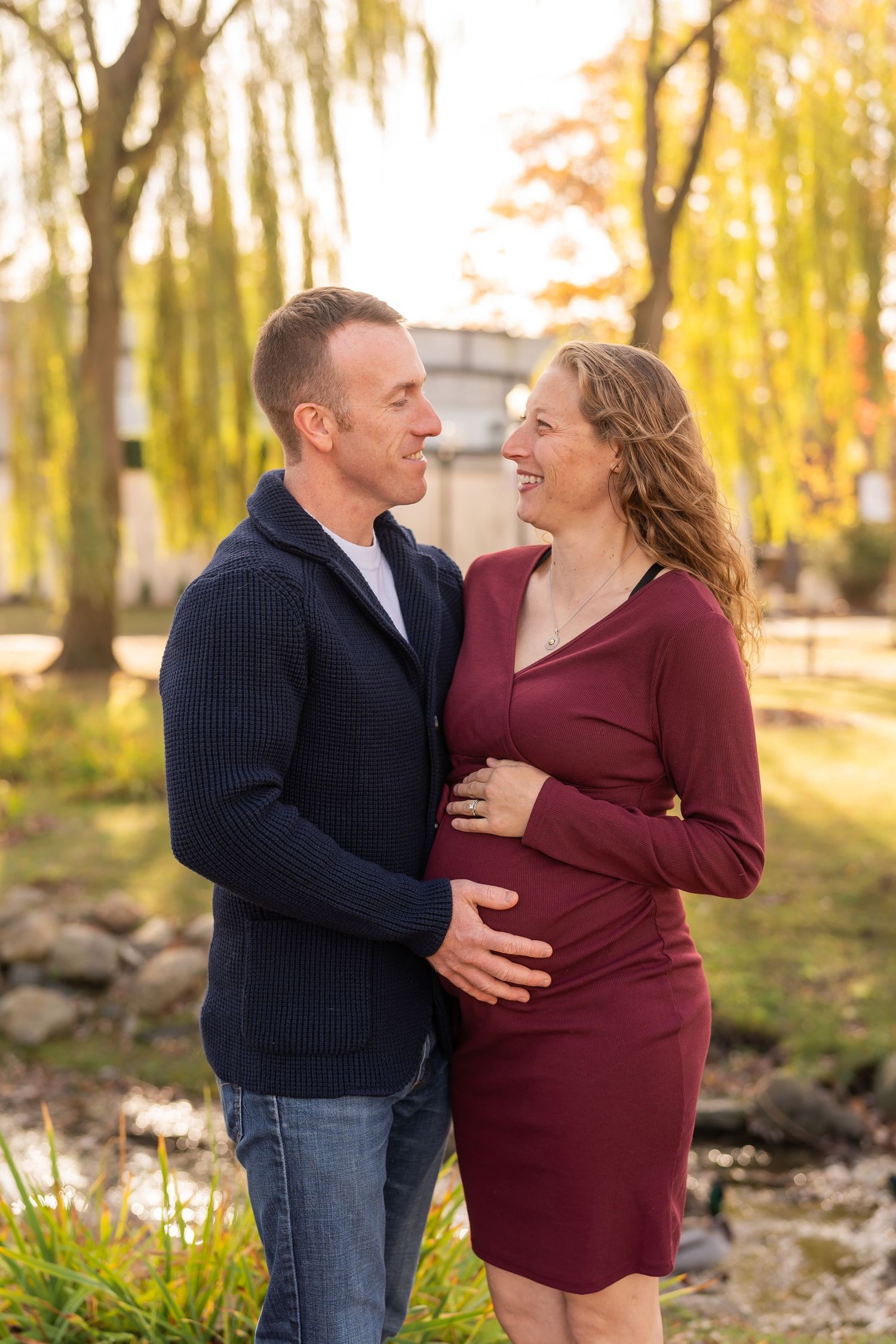 A couple shares a tender moment outdoors during an autumn maternity photo session with golden willows in the background.