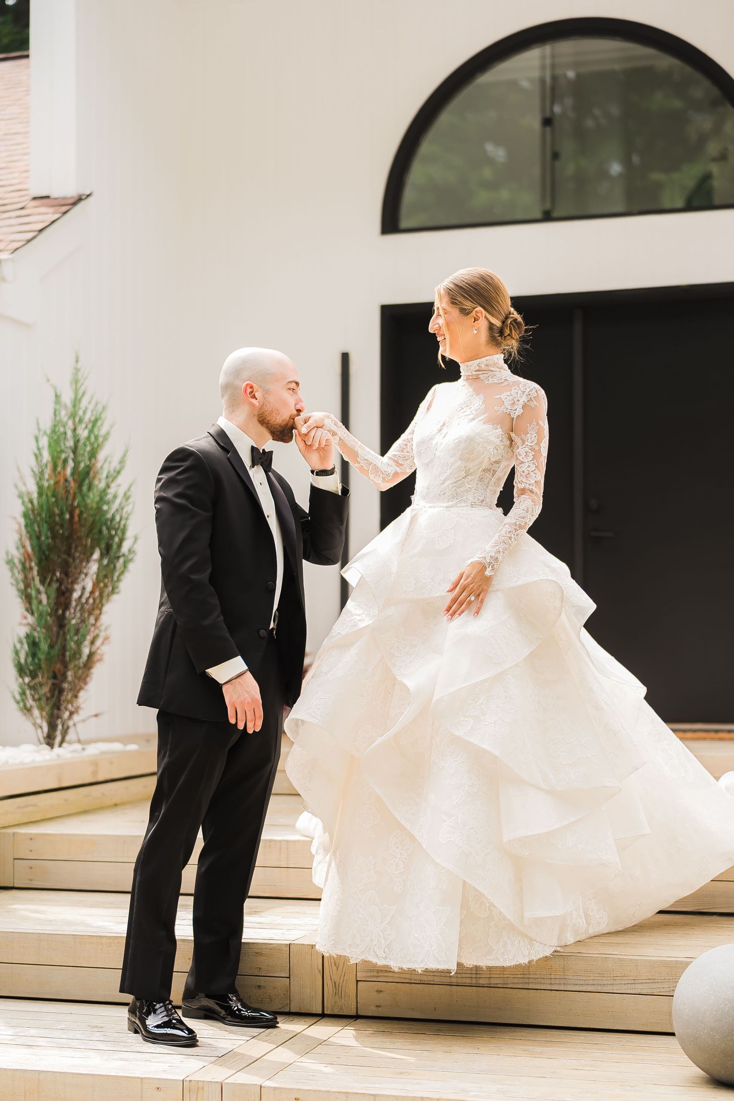 Elegant couple on steps as groom kisses bride's hand, her layered white gown flowing beautifully.