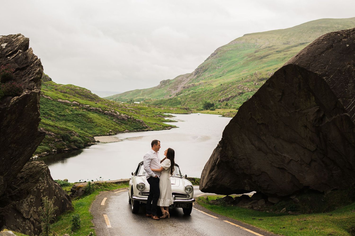 Newlyweds sit on vintage car trunk surrounded by dramatic mountain landscape on misty road.